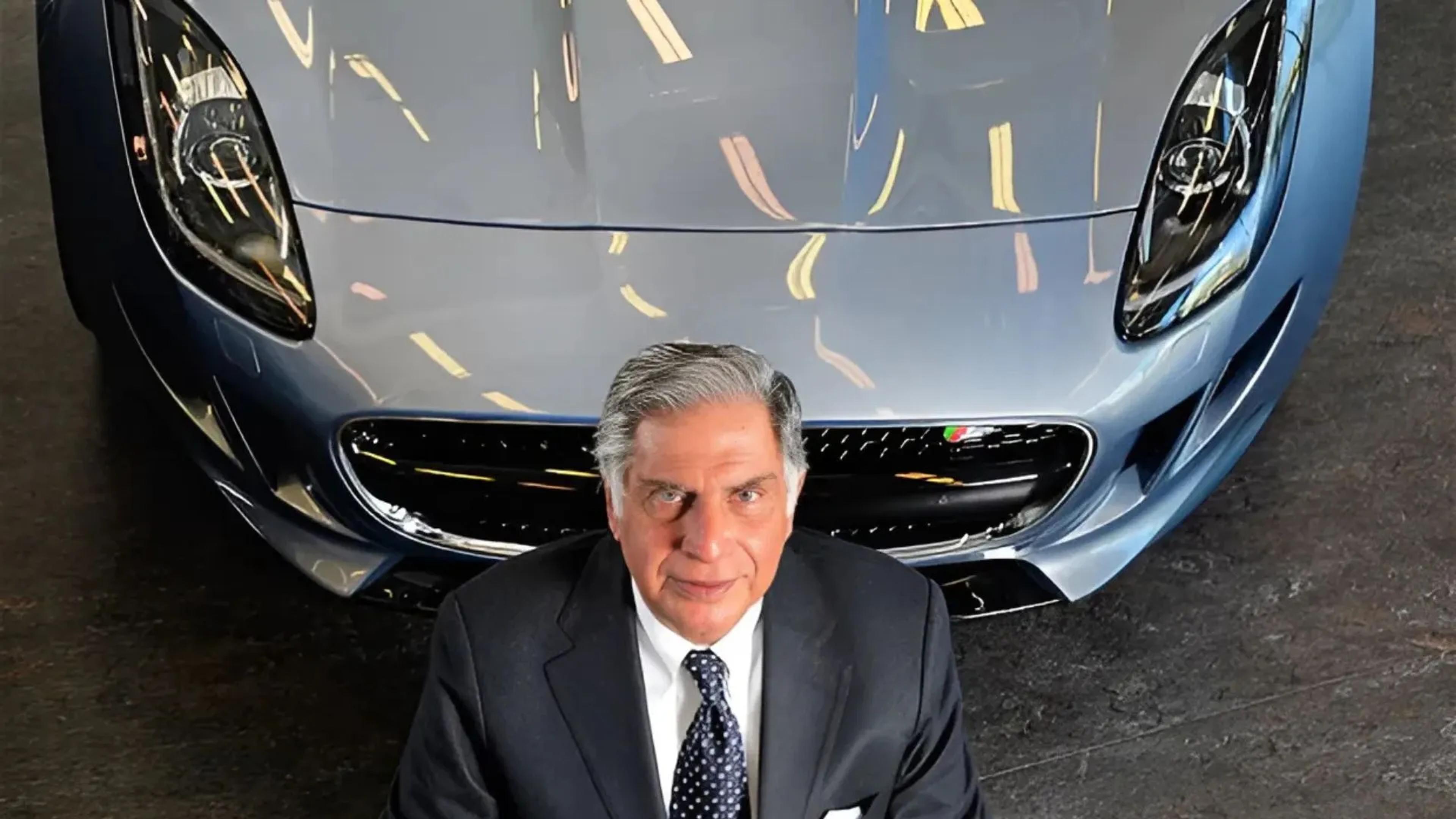 The image shows Ratan Tata, the distinguished former chairman of Tata Sons, standing in front of a luxurious car, likely a Jaguar, showcasing his influence in the automotive industry. He appears confident and composed, wearing a dark suit with a tie, with a subtle smile. The setting suggests a showroom or a similar indoor venue, with reflections of lights on the car's shiny surface.