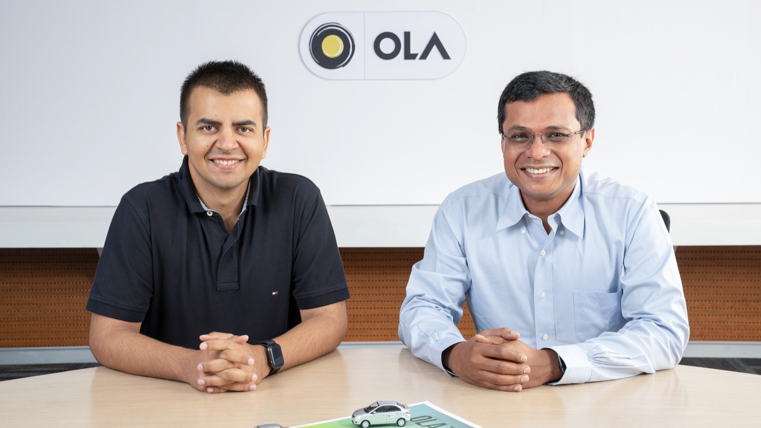 Bhavish Aggarwal, CEO of Ola, and Sachin Bansal smiling together at a conference table with Ola branding in the background, signifying their professional partnership and investment
