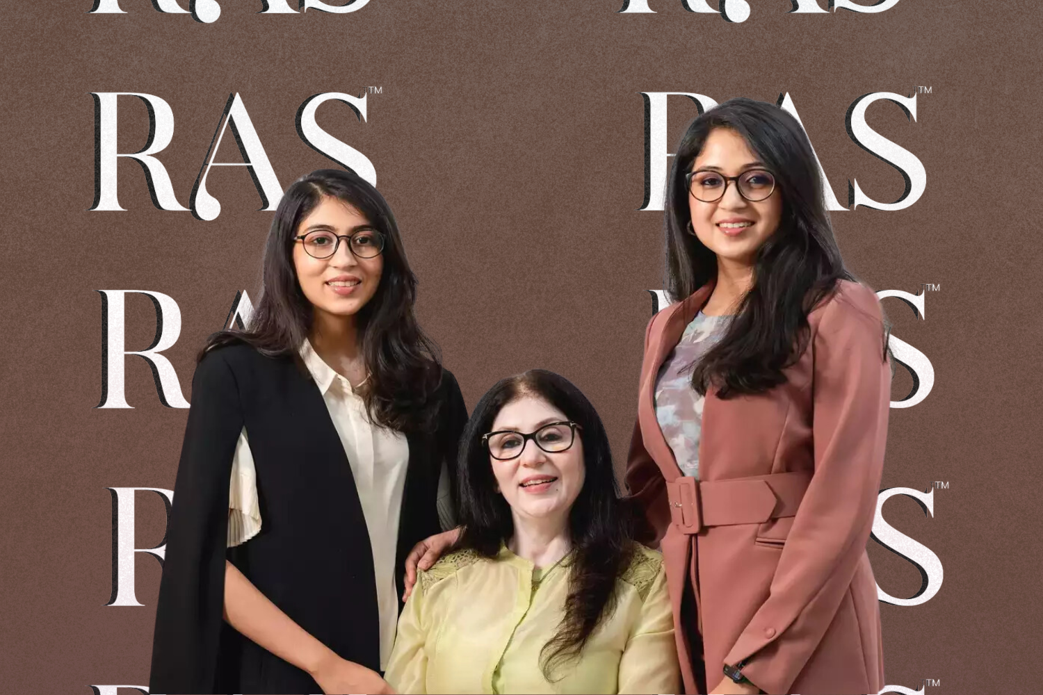 Three women standing together against a brown background with the "RAS" logo repeatedly visible. The women are dressed in formal attire, with one seated in the middle and the others standing on either side, all smiling and wearing glasses.