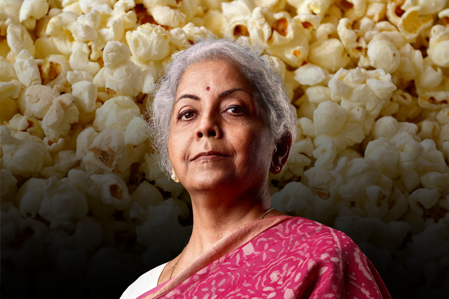 A confident older woman in a pink sari with white patterns stands in front of a background filled with popcorn. The image has a subtle gradient darkening effect toward the bottom.