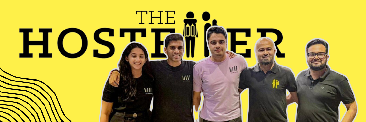 Group photo of five team members standing together in front of The Hosteller logo on a bright yellow background. Two men and one woman from V3 Ventures are seen wearing black shirts, while two men from The Hosteller wear casual T-shirts, including one with the brand's logo. Everyone is smiling and standing closely, reflecting a sense of collaboration and partnership.