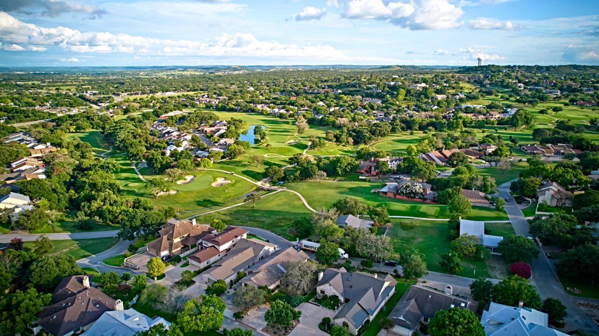 Aerial view of an upscale residential community featuring lush golf courses, winding paths, a small lake, and sprawling homes under a bright blue sky. Houston, Texas, USA