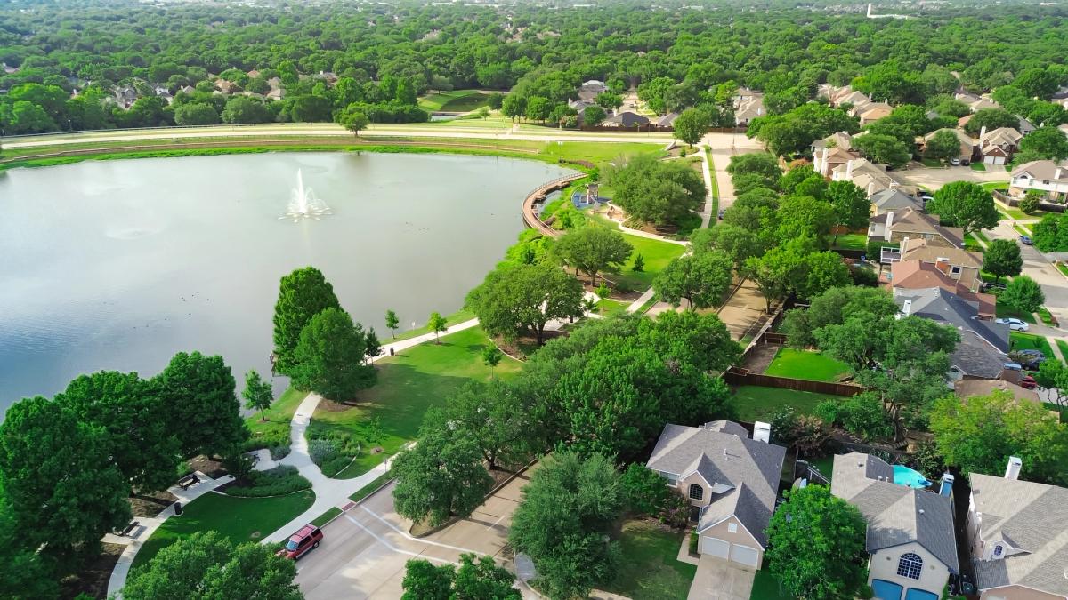 Lush green community Rheudasil Park in Flower Mound, Texas with curved boardwalk, walking trails, playground, upscale suburban two-story houses with resort-style swimming pool, grassy lawn, aerial