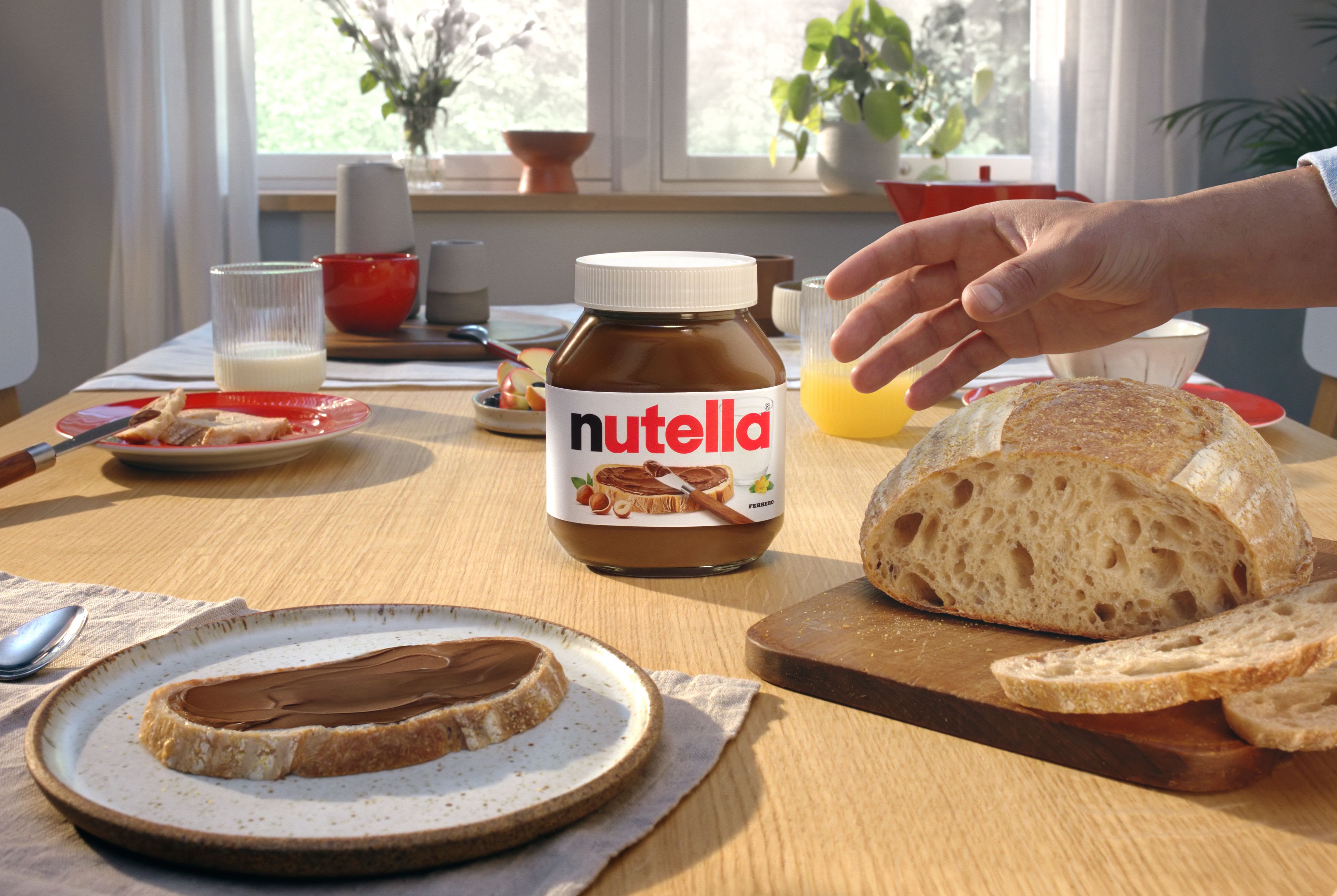 En burk Nutella® placerad på ett frukostbord med en skiva bröd med Nutella®-pålägg brevid. I bilden syns även färskt bröd, en hand som sträcker sig mot bordet, och olika frukosttillbehör som juice och mjölk. Solen skiner in genom fönstret och skapar en ljus och inbjudande atmosfär