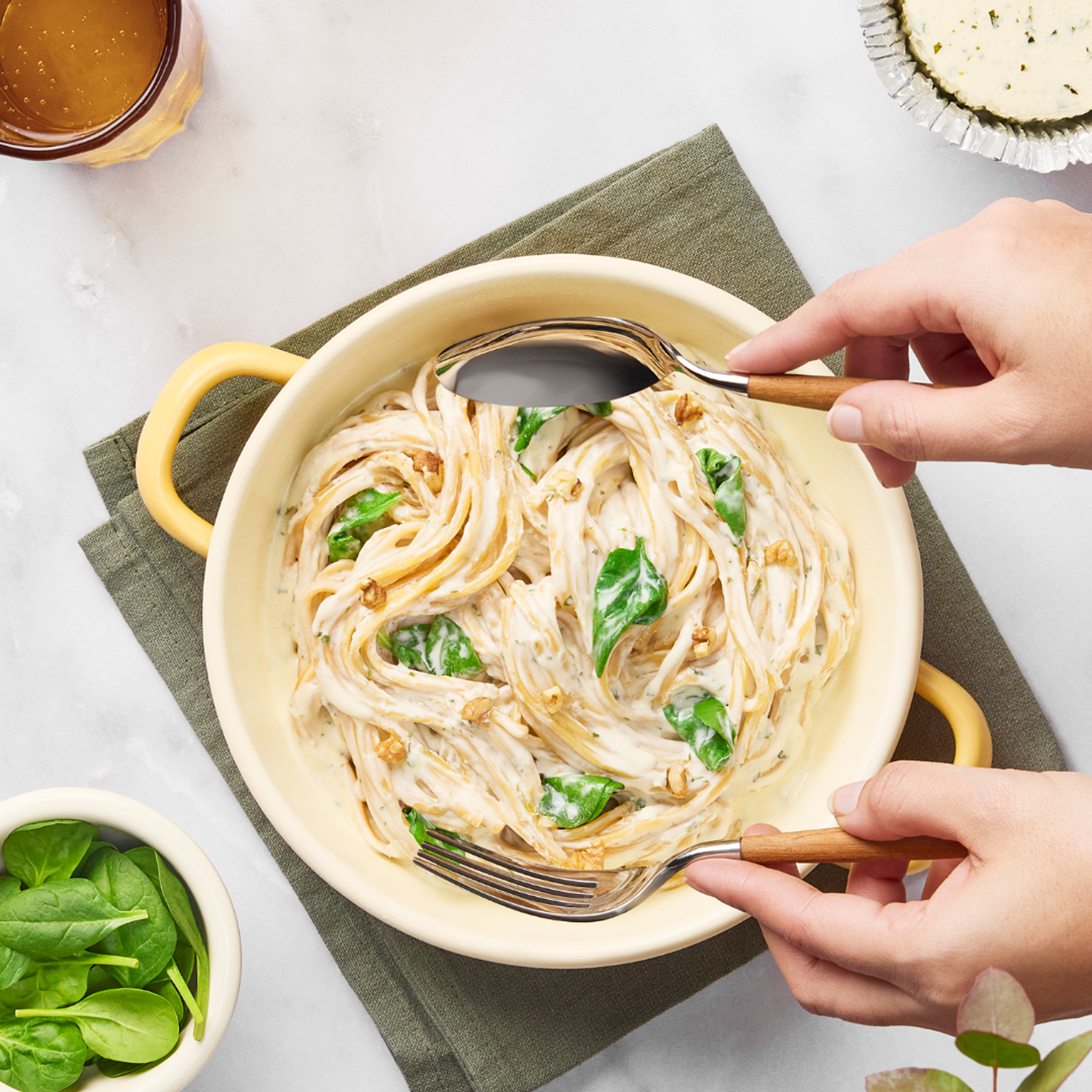 Linguine pastasallad blandad med en krämig Boursin-sås och färsk spenat i en gul gryta. Två händer blandar pastan med en sked och gaffel.