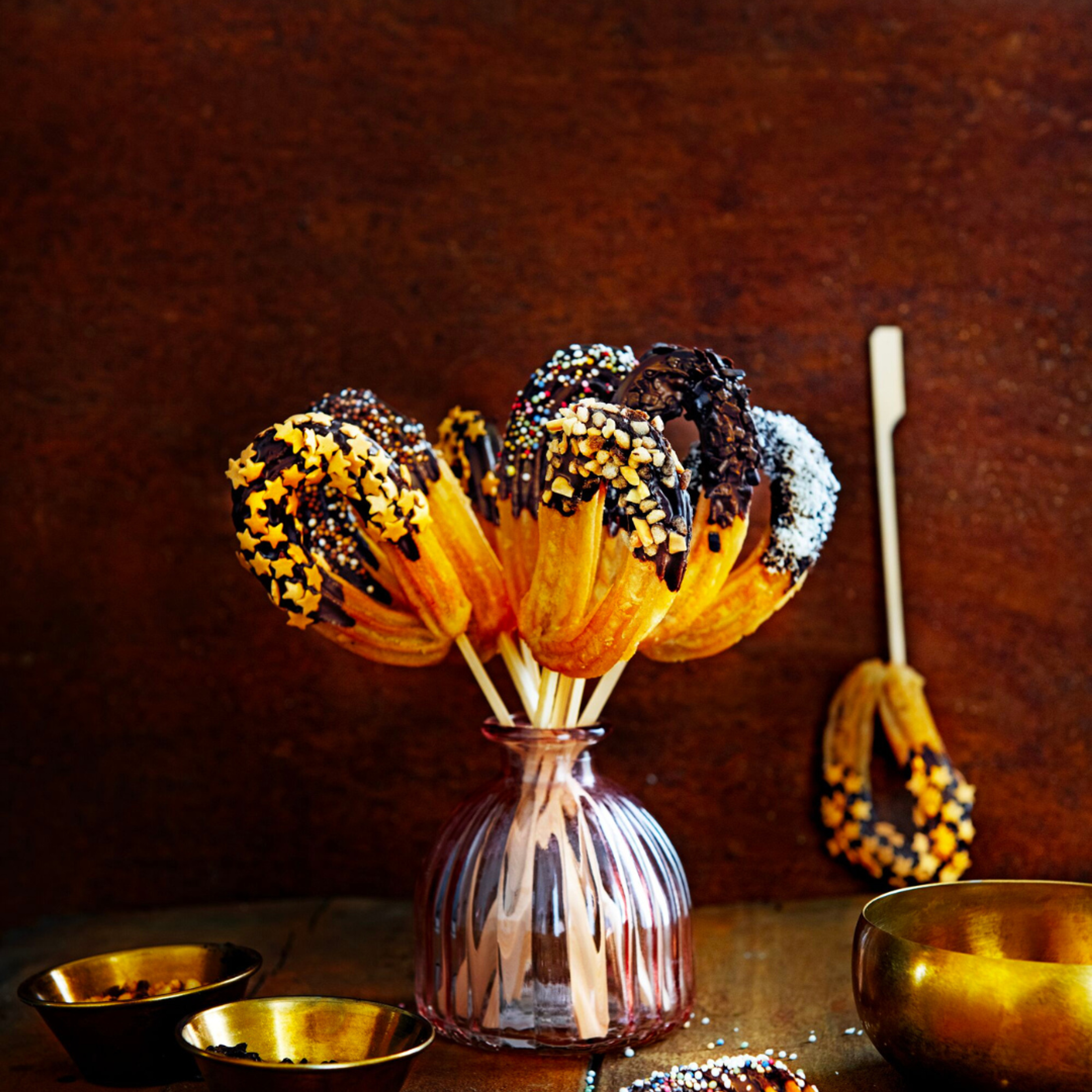 Churros på pinnar doppade i choklad och dekorerade med strössel, stjärnor och socker, arrangerade i en glasvas på ett bord.