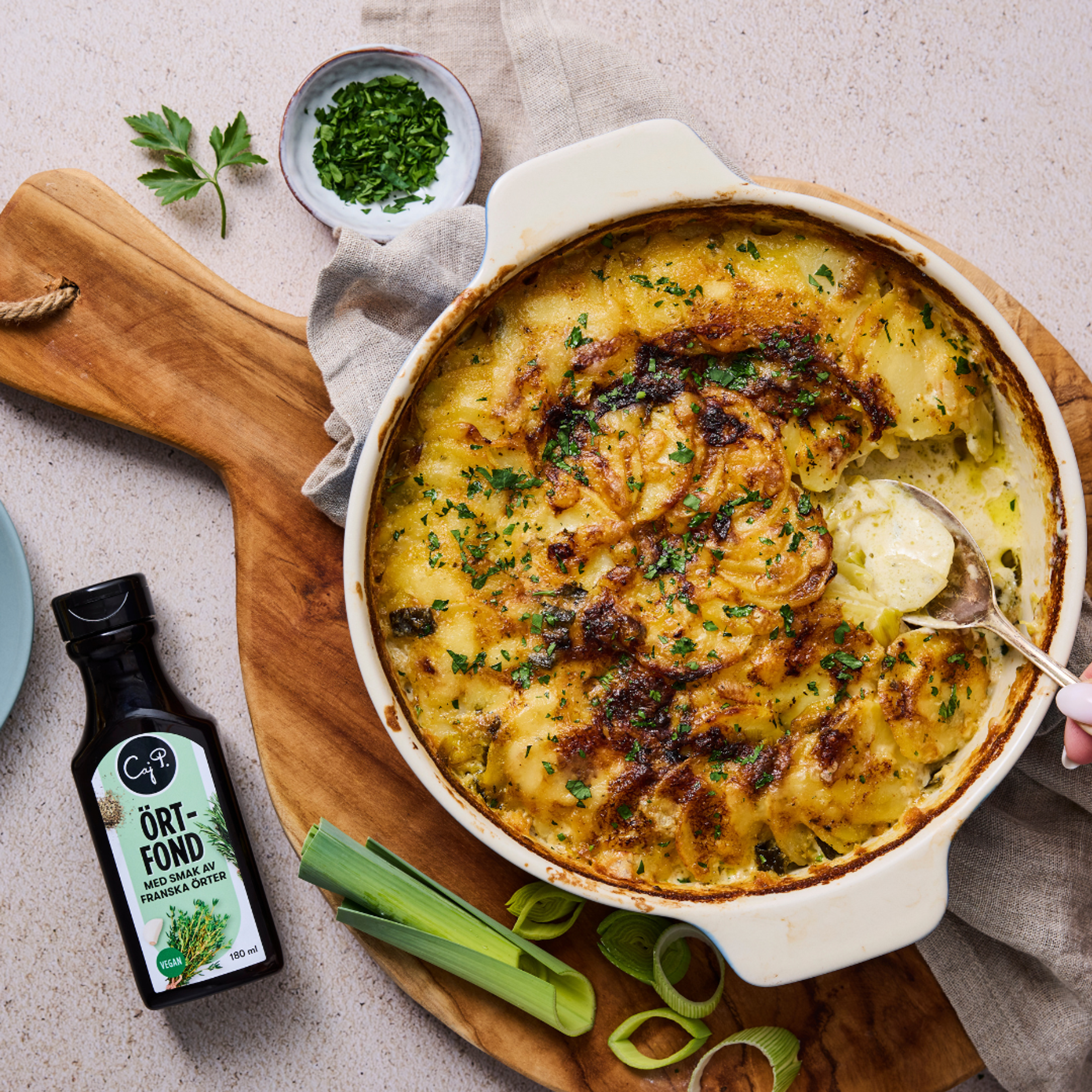 Krämig potatisgratäng med purjolök i en ugnsform, toppad med persilja och gratinerad yta. Bredvid ligger en flaska Caj P’s örtfond samt purjolök och persilja på ett träfat.