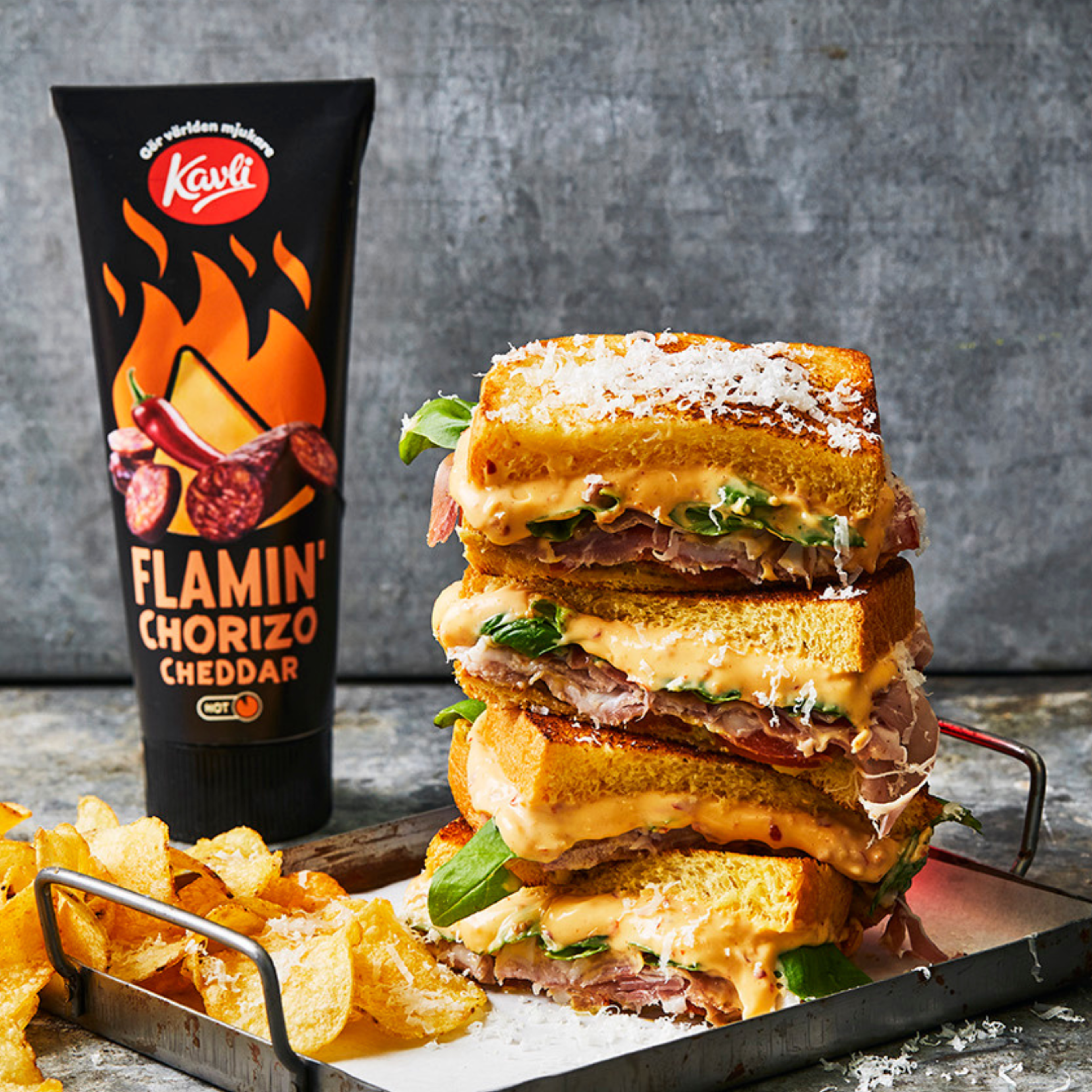 En hög med varma, ostiga mackor med prosciutto på en bricka, med chips bredvid och en tub Kavli Flamin’ Chorizo Cheddar i bakgrunden.