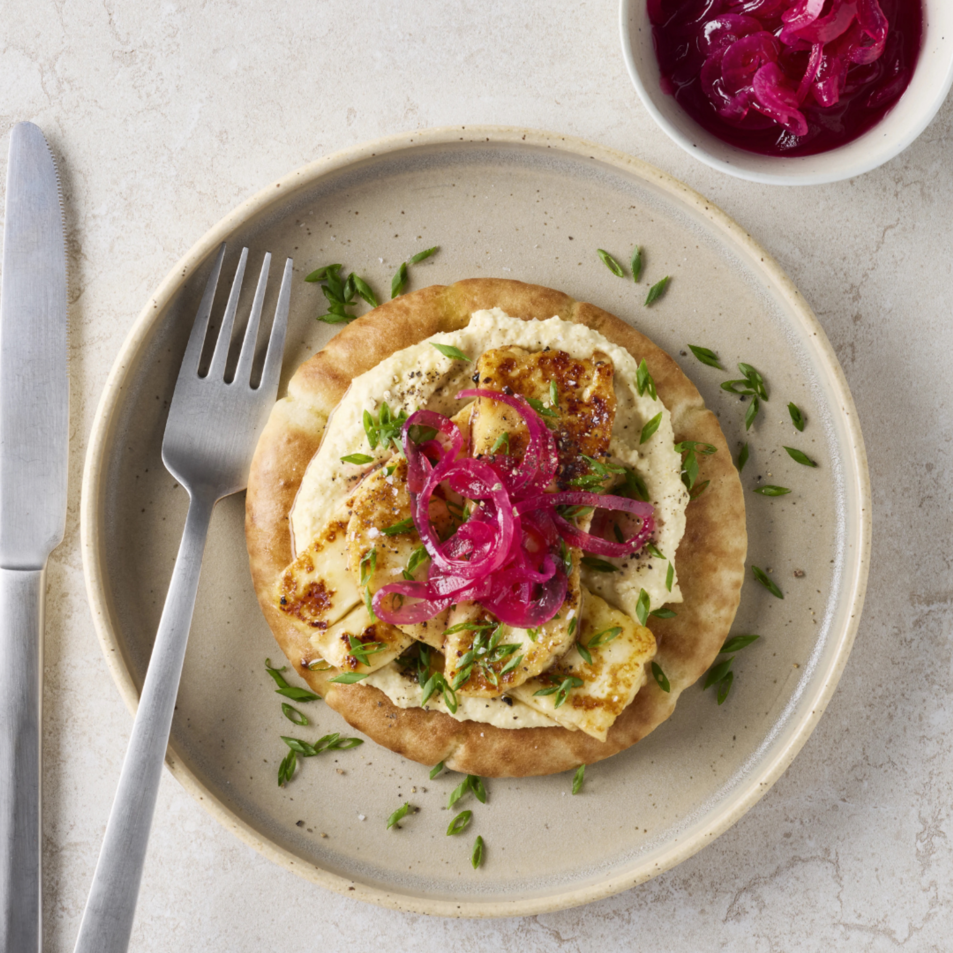 Pitabröd toppat med hummus, honungsglacerad tryffelhalloumi, picklad rödlök och finhackad gräslök serverat på en tallrik.