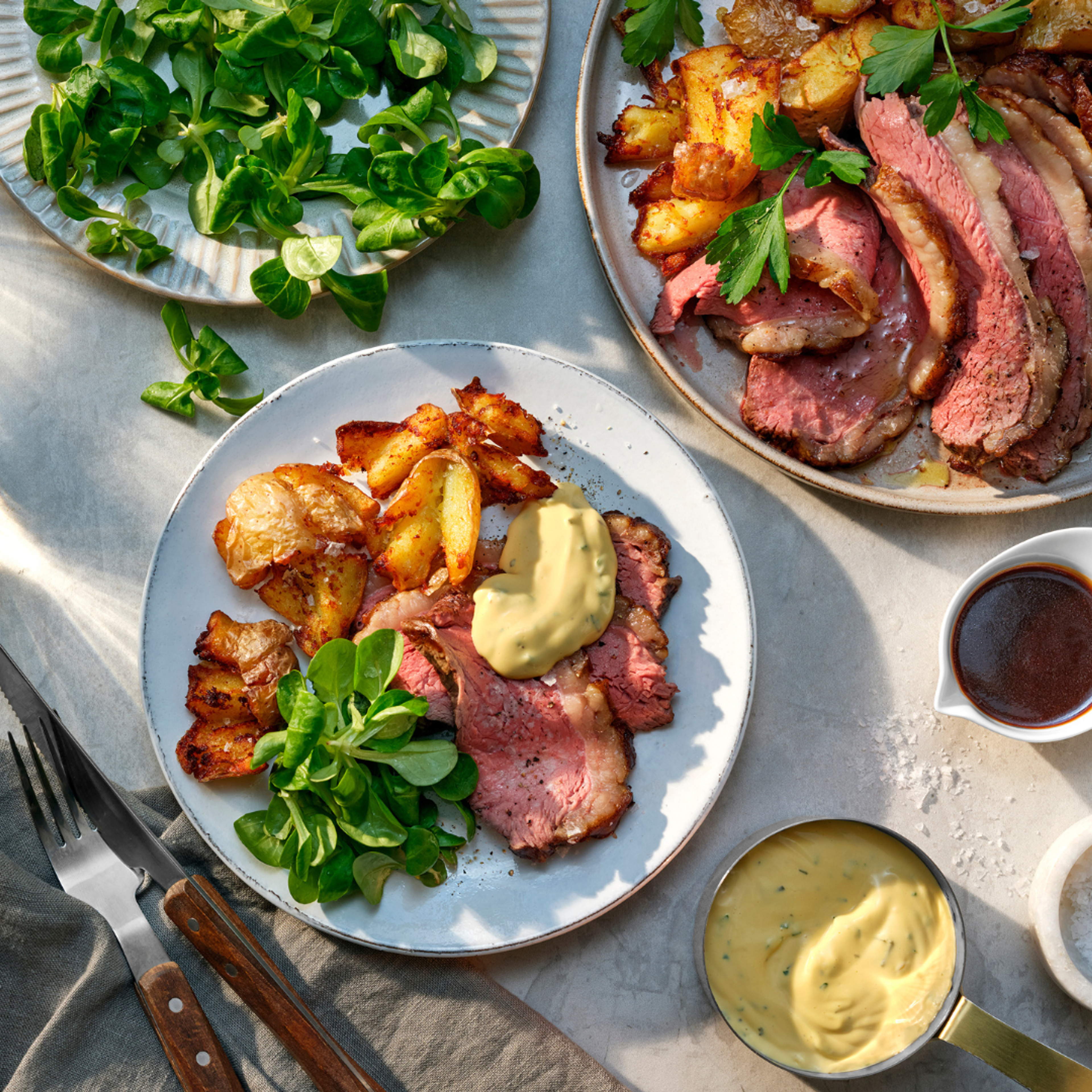 Tallrik med skivad grillad pichana, friterad smashed potatis, machésallad och en klick Lohmanders Bearnaise. Runtom syns fler tallrikar med kött och potatis samt en kastrull med varm bearnaisesås från Lohmanders.