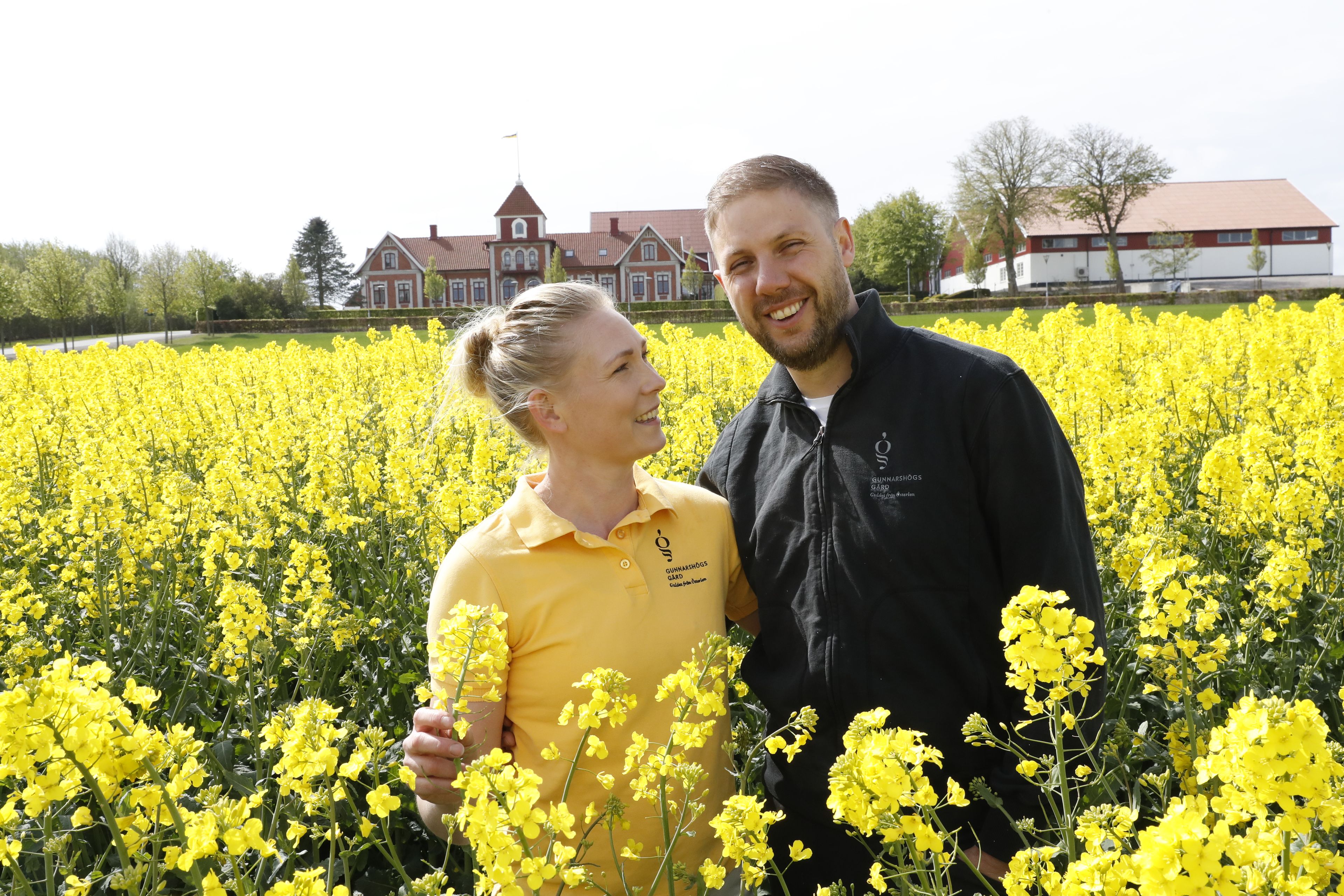 Anja och Erik Persson från Gunnarshögs Gård står i ett fält av blommande raps på Österlen, med gårdens byggnader i bakgrunden