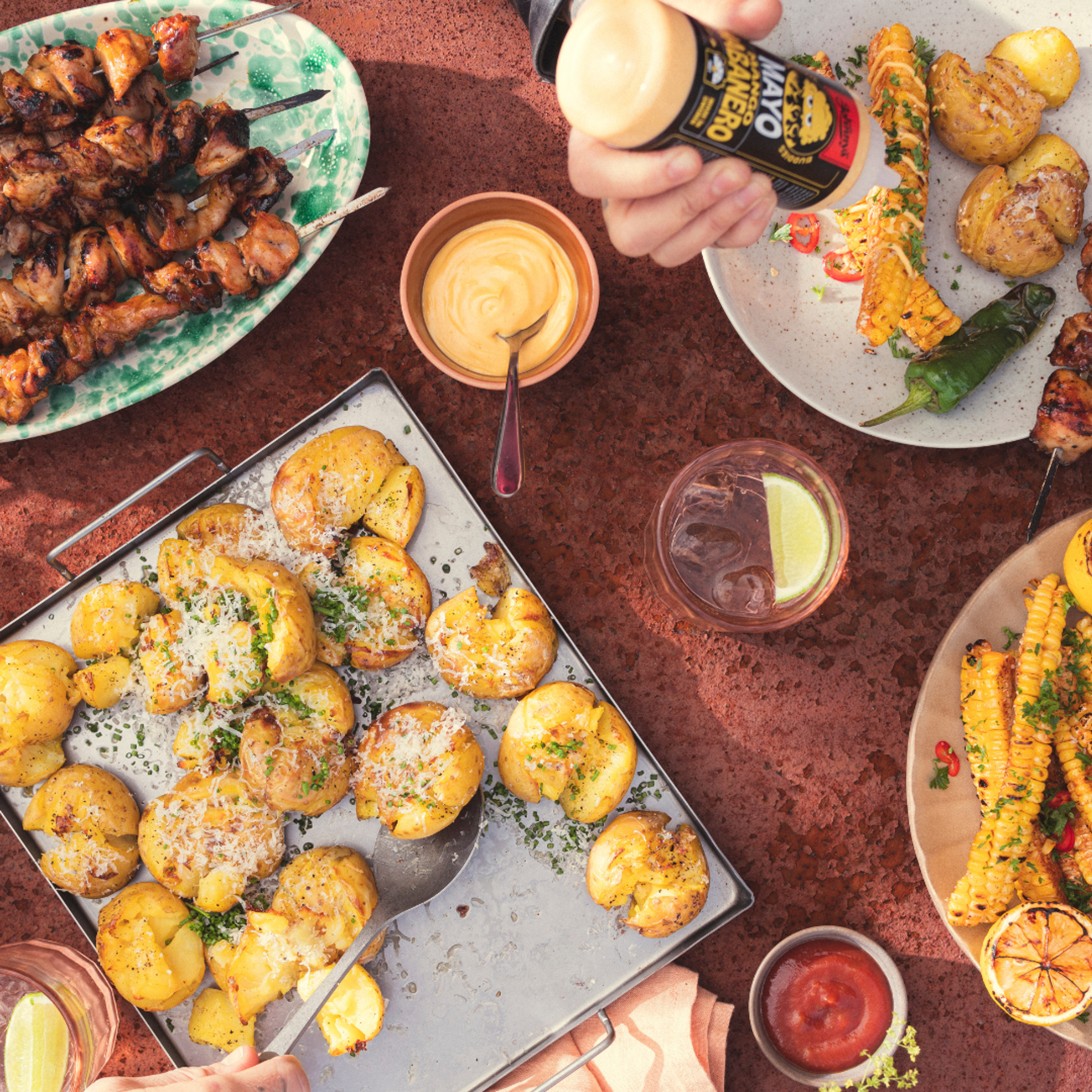 Grillbord med kycklingspett, smashed potatoes toppade med parmesan, grillade corn ribs och en skål Mango Habanero-mayo. En hand håller en flaska Johnny’s Mayo Mango Habanero ovanför maten.