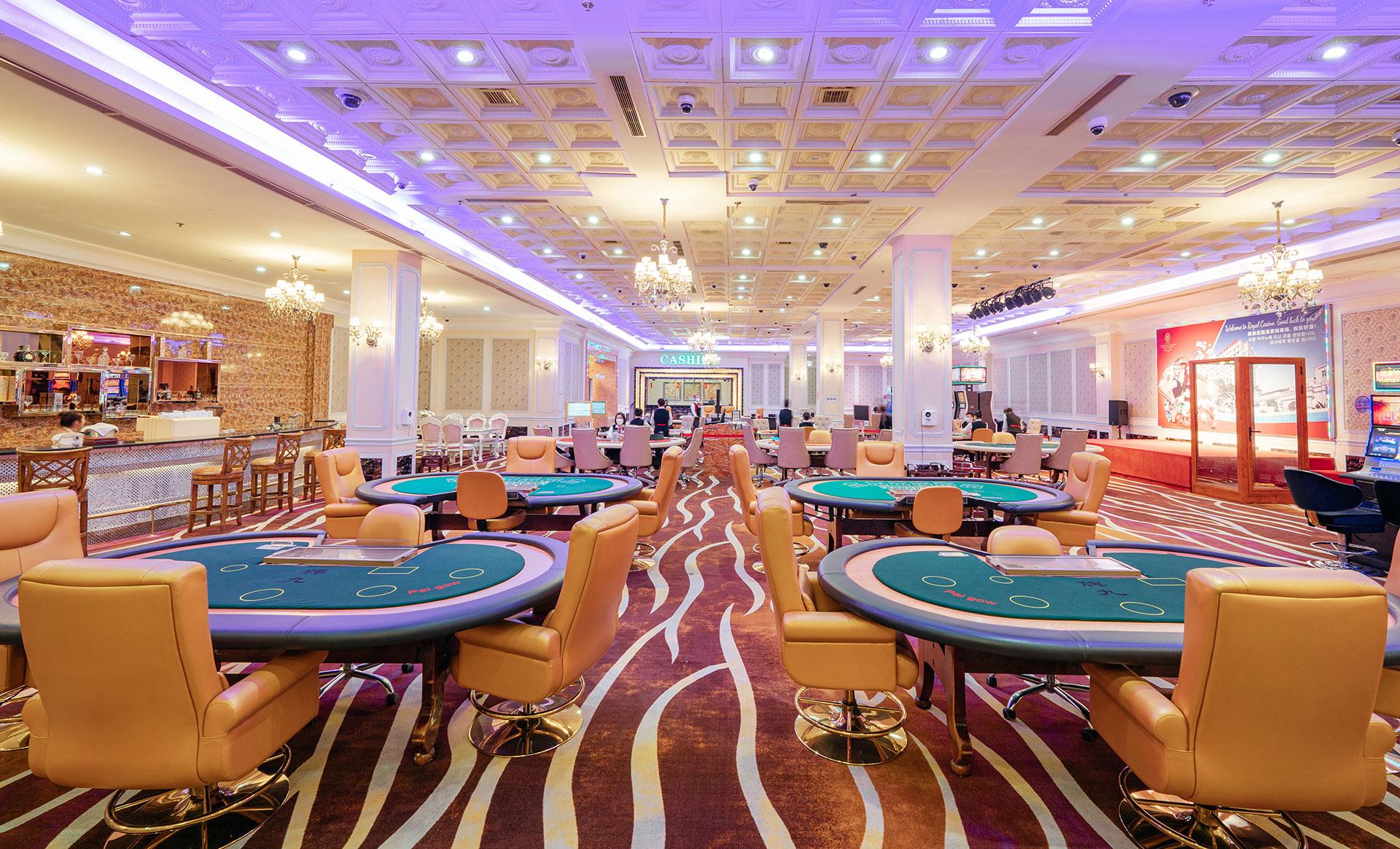 Sảnh casino với các bàn chơi mặt nỉ xanh viền gỗ và ghế da màu bò, trần ô kẻ gắn đèn chùm pha lê, thảm hoa văn sóng, quầy bar ốp đá bên trái và quầy thu ngân ở cuối sảnh