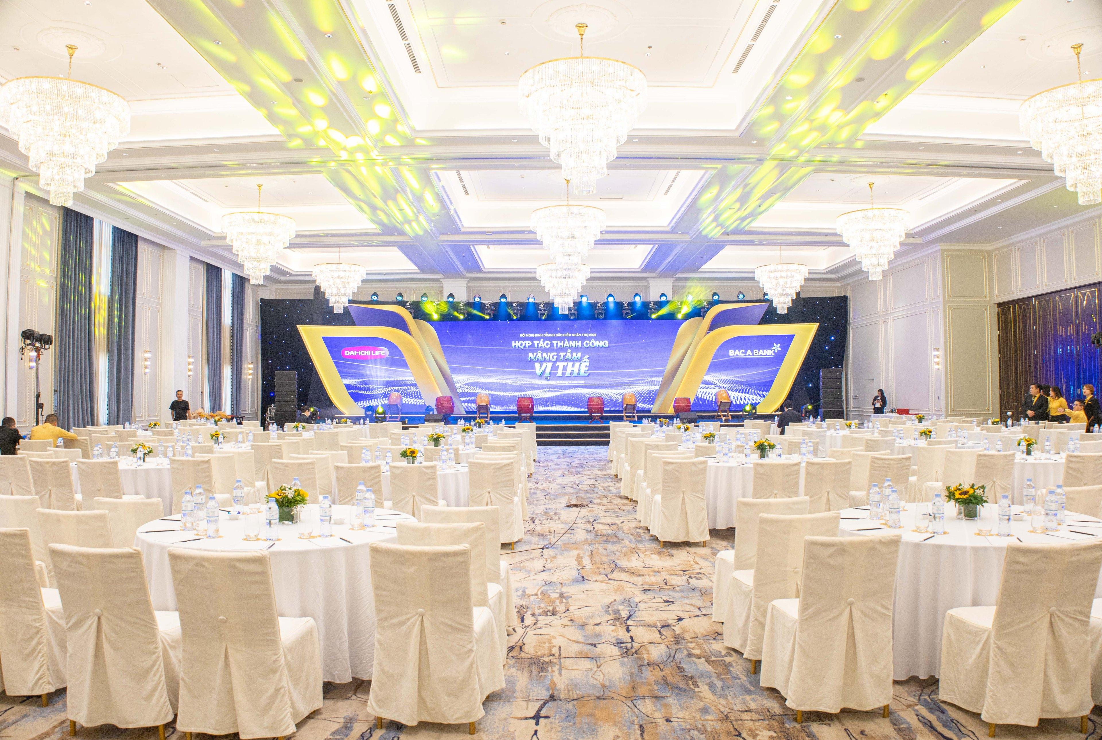 A ballroom laid for a banquet: dozens of round tables in white linen facing an LED stage, crystal chandeliers down both sides and yellow-and-blue stage lights washing the ceiling