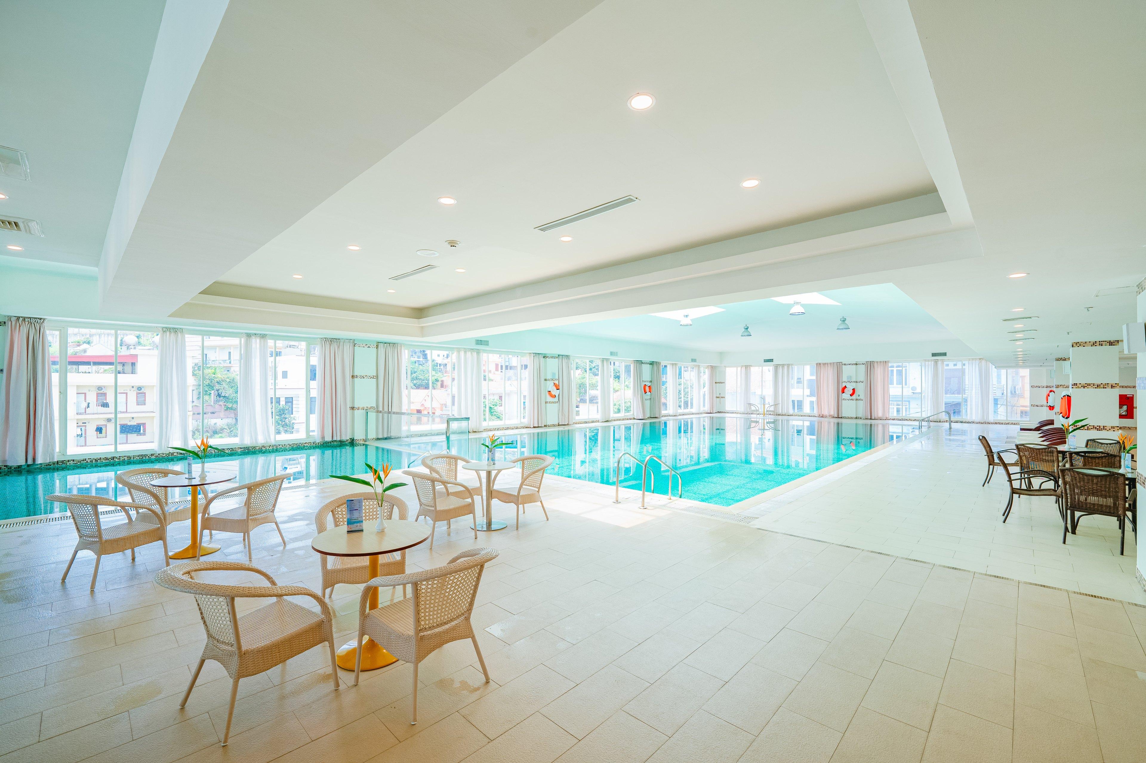 Round rattan tables and chairs set in groups along the indoor poolside, a stepped ceiling above