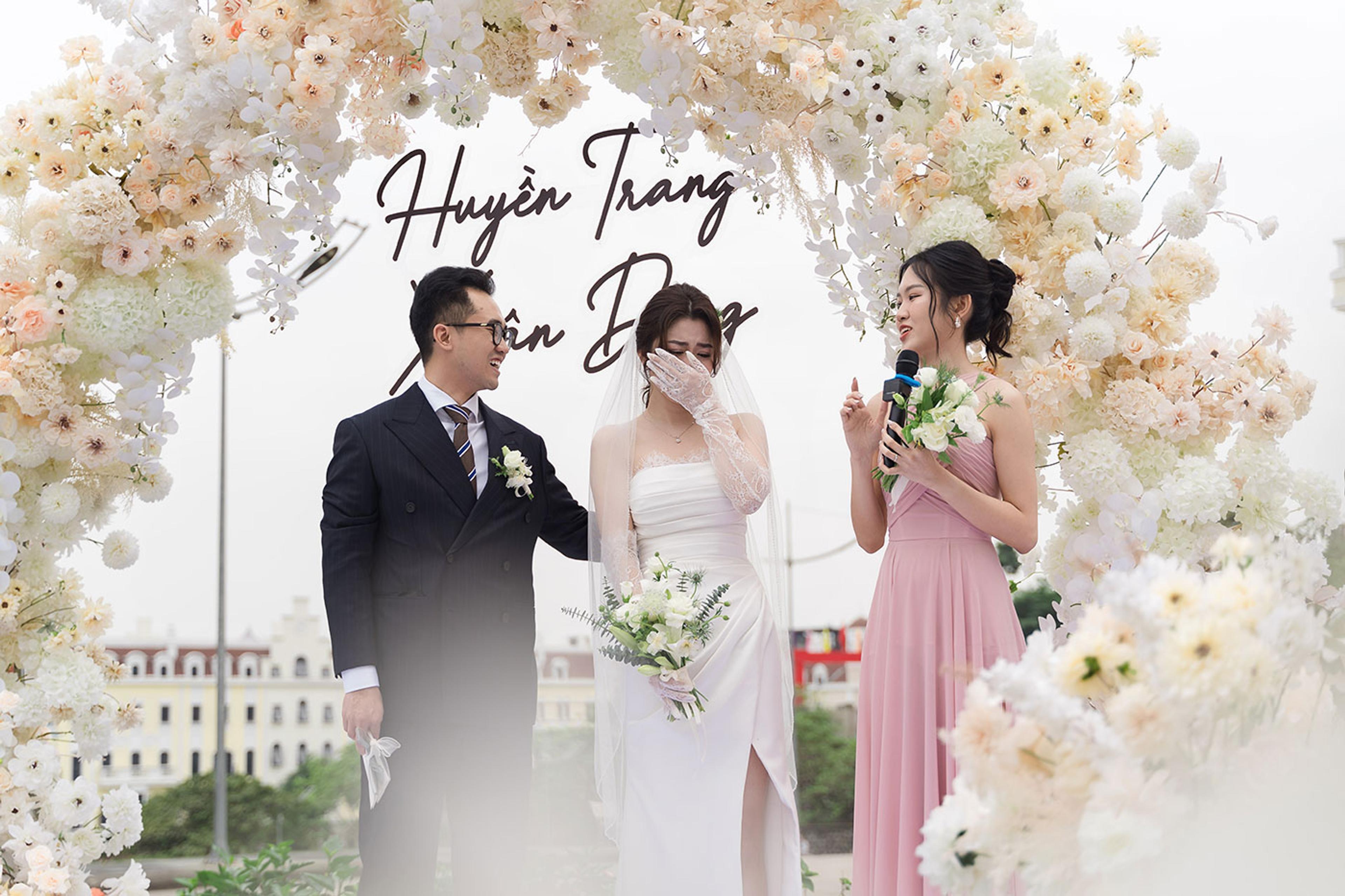 A bride and groom beneath a white flower arch, a host with a microphone beside them