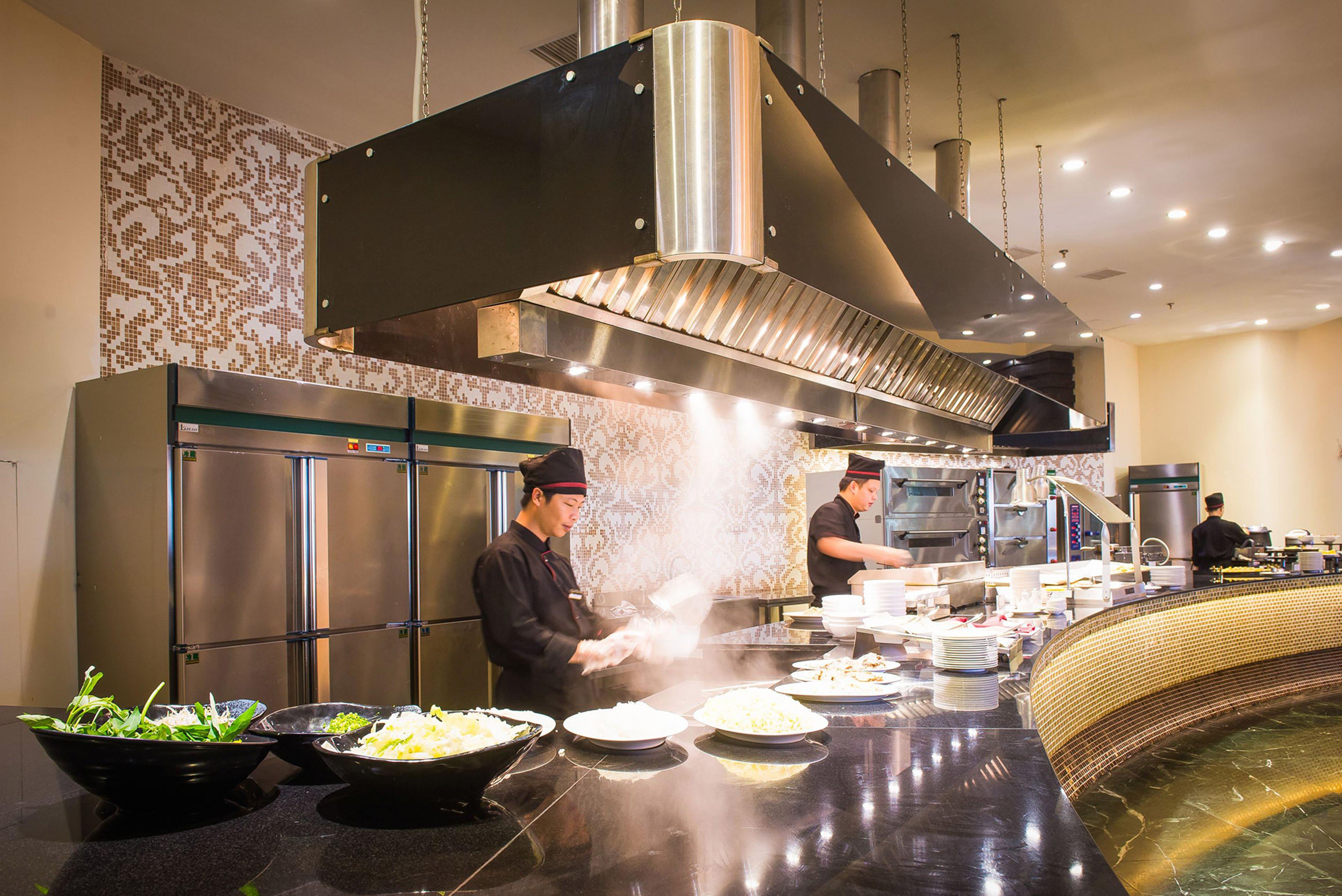 Chefs cooking behind an open kitchen counter under a large extractor hood, steam rising over the buffet line