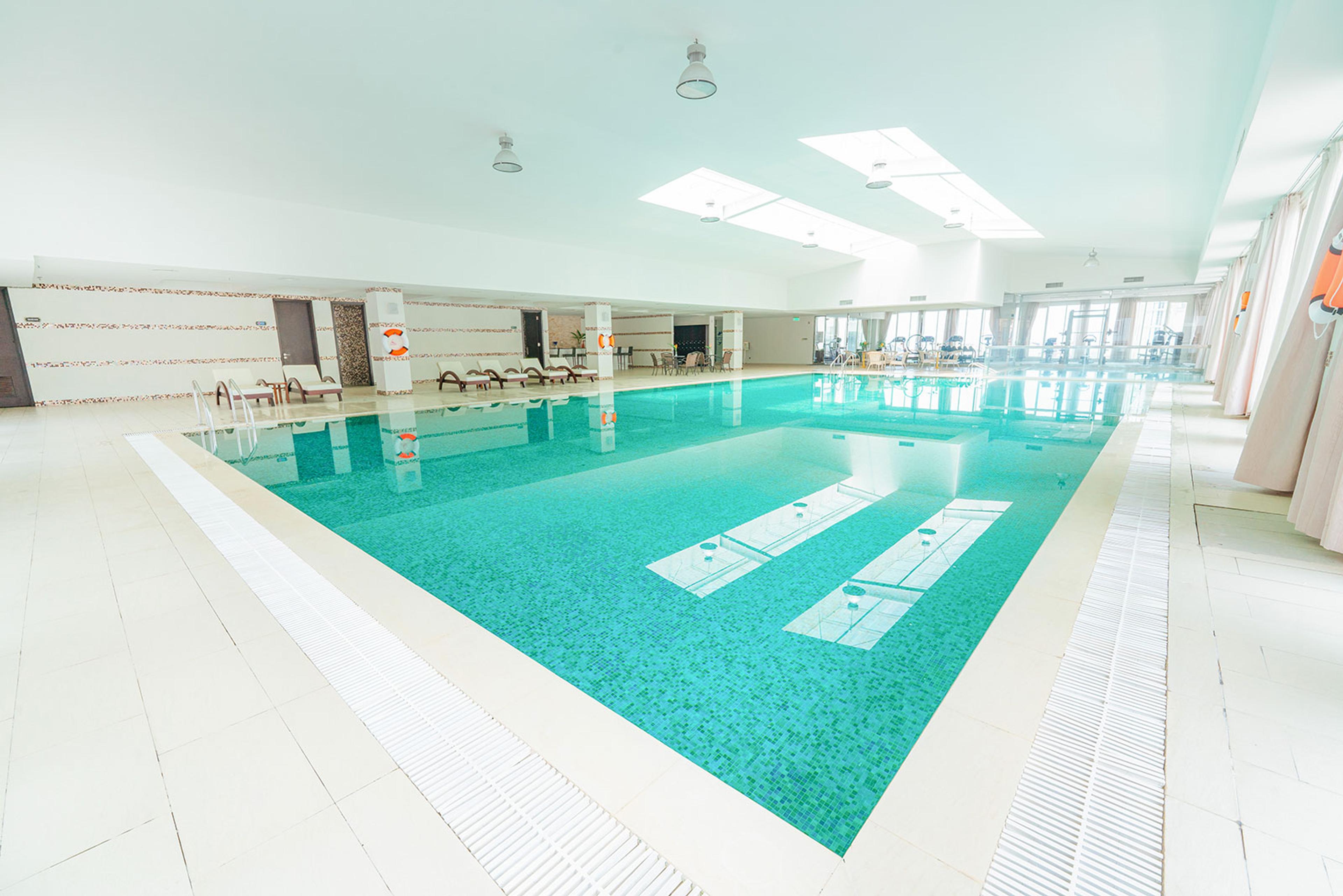 ターコイズ色の水をたたえた屋内プール。デッキと周囲は白いタイル張りで、頭上には採光用の天窓があります