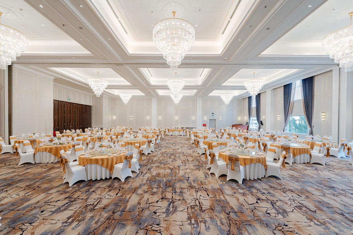 Ha Long Ball Room ที่จัดโต๊ะจัดเลี้ยง โต๊ะกลมคลุมผ้าสีทองบรอนซ์กับเก้าอี้คลุมผ้าขาว โคมระย้าคริสตัลเรียงแถวใต้เพดานหลุม และพรมลายหินอ่อนปูเต็มพื้น