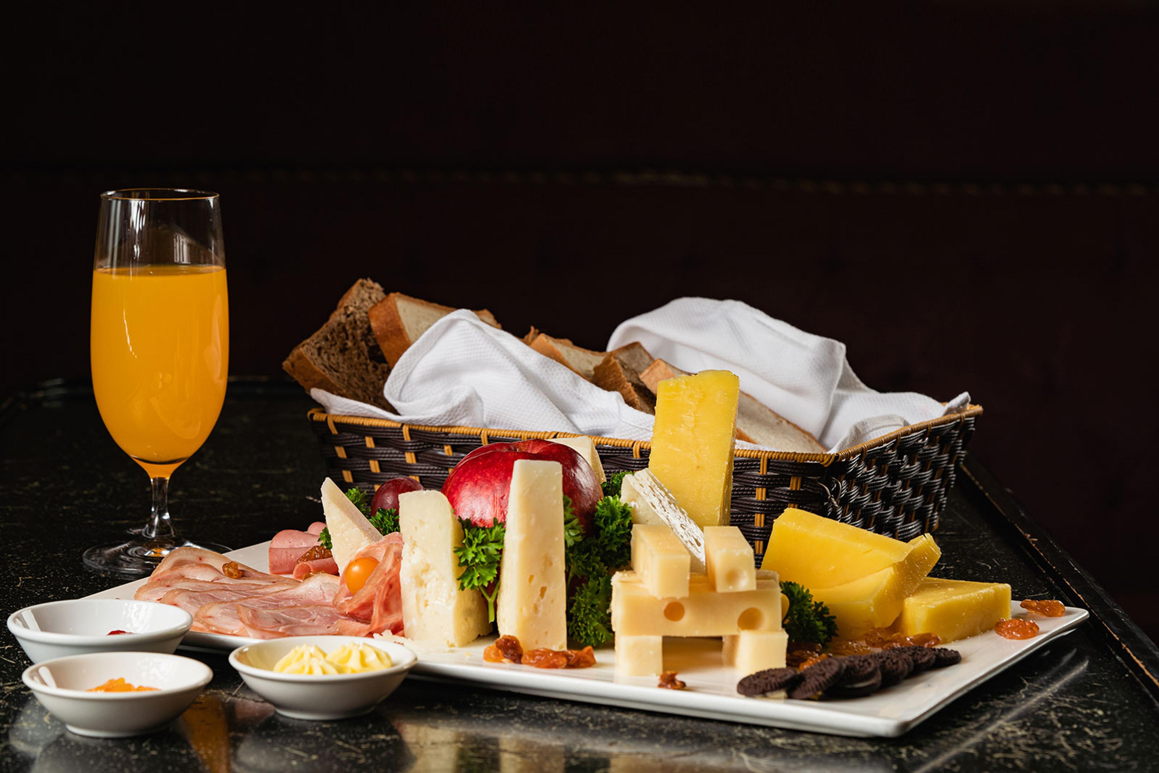 A platter of cut cheeses, cured meats, a bread basket and a glass of orange juice on a dark surface