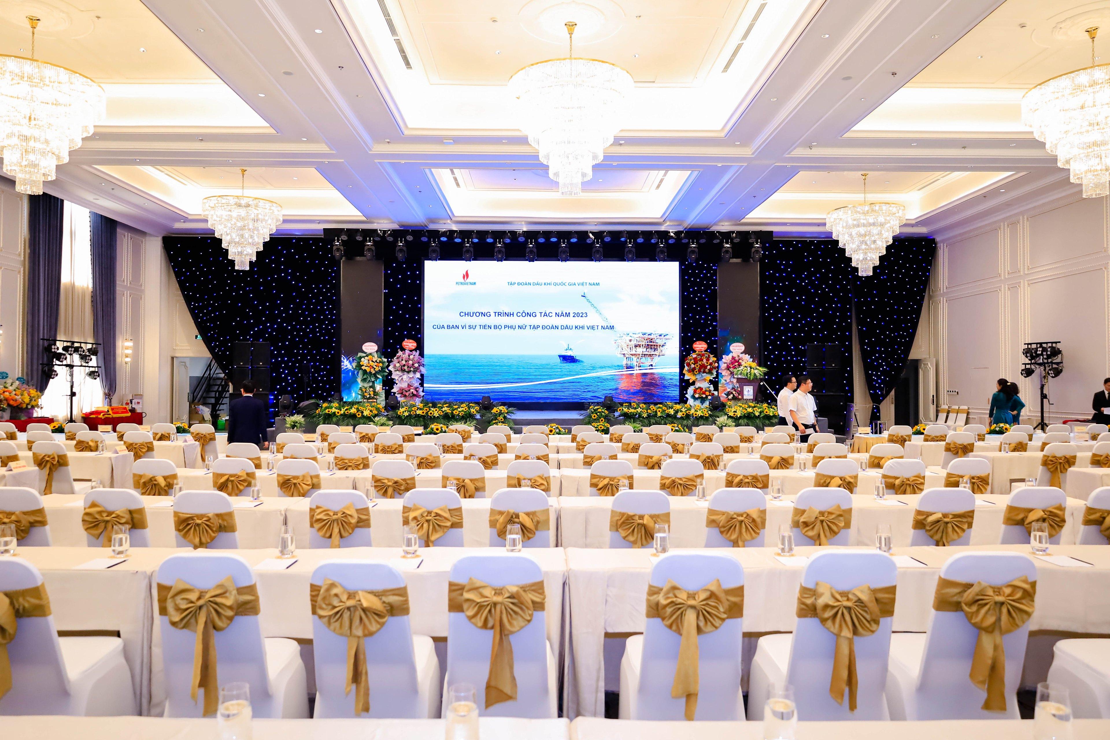 A hall in theatre layout: hundreds of white chair covers with gold sashes facing an LED stage, a black star-cloth backdrop behind it and crystal chandeliers down both sides