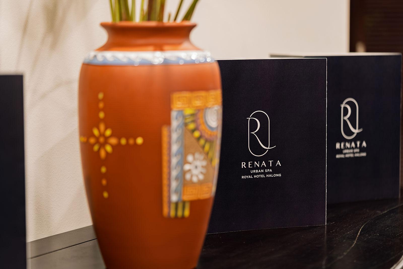 R のロゴが入った紺色の Renata Spa のパンフレットが濃い色の木製台に立てられ、手前には模様を描いたテラコッタの花瓶が置かれています