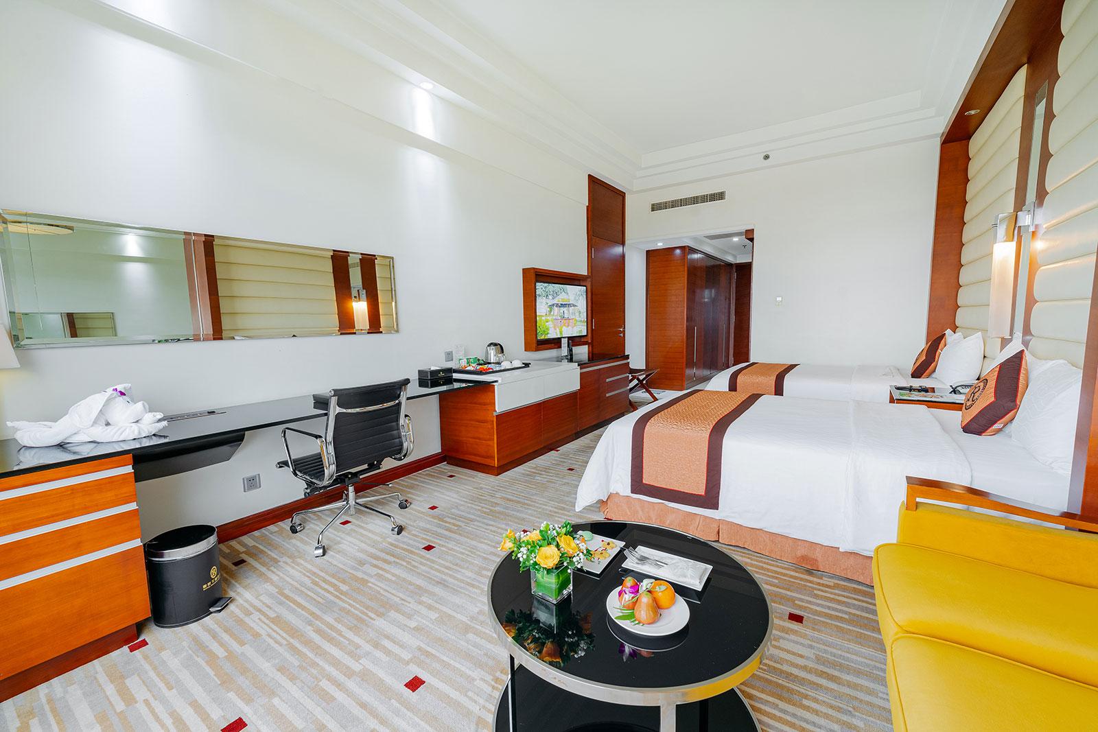 The work corner of a Deluxe room: a long desk with a swivel chair and a round table set with flowers and a plate of fruit, the two beds behind
