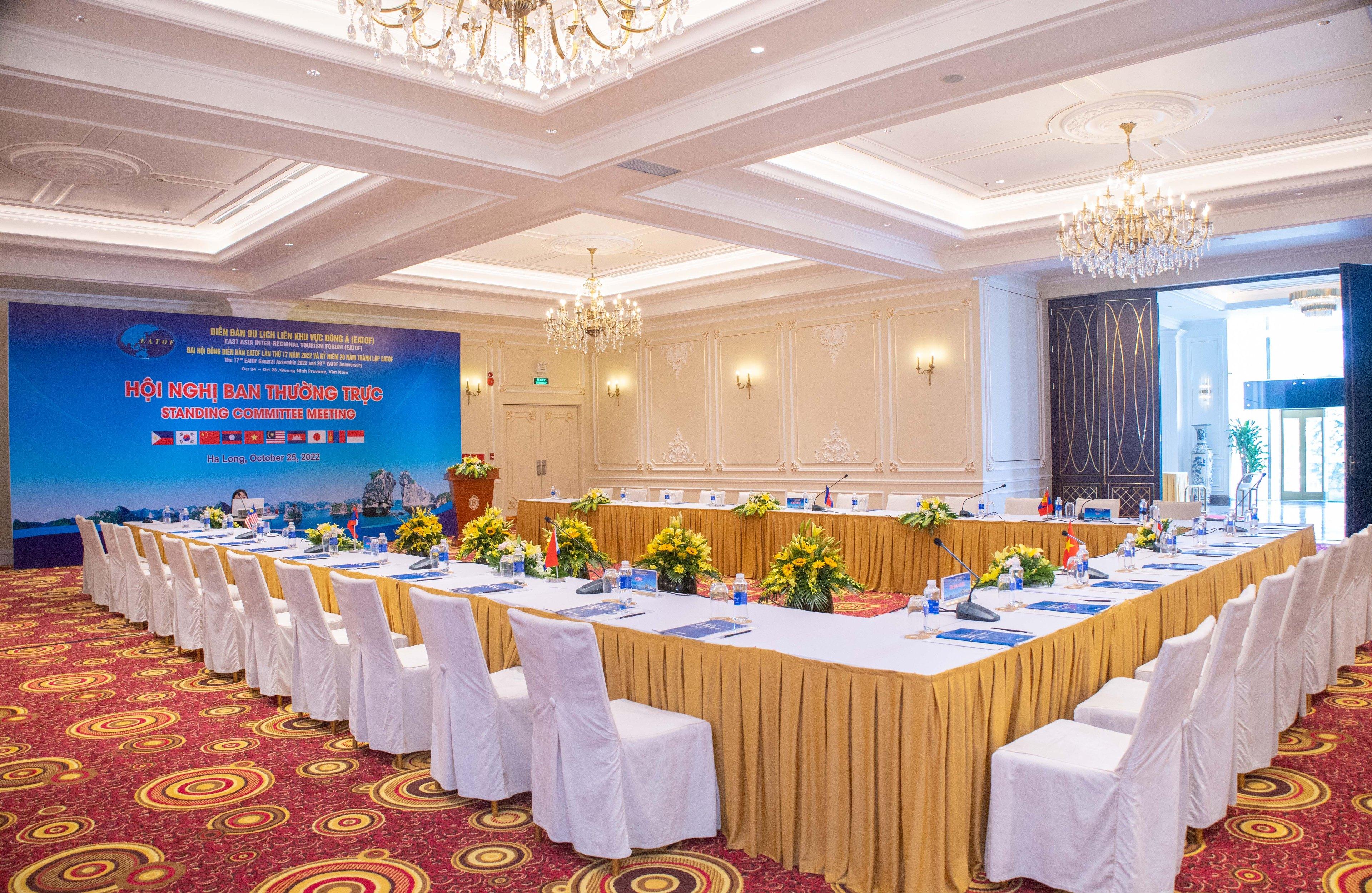 A meeting room in hollow-square layout: tables skirted in gold joined into a large rectangle, white-covered chairs around them, and a conference backdrop on the screen at the head of the room