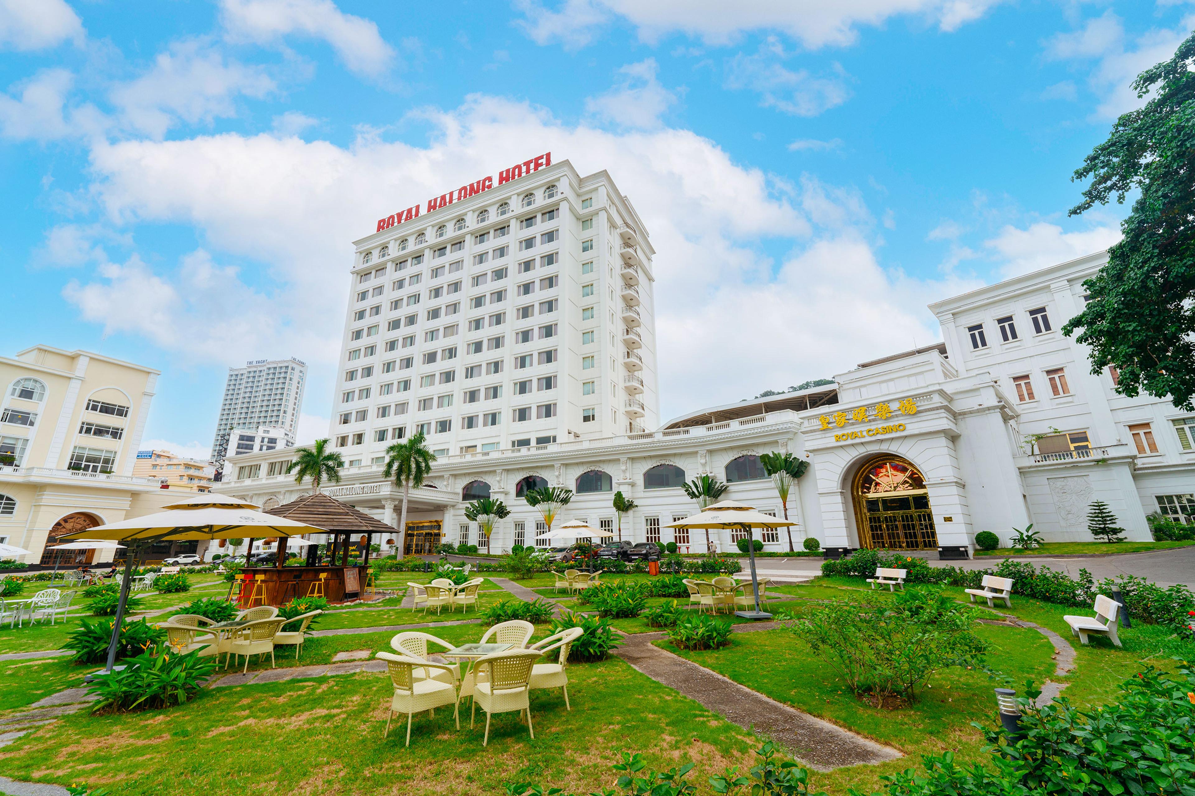 Toà khách sạn màu trắng với biển hiệu ROYAL HALONG HOTEL trên nóc, nhìn từ khu vườn có ô che và bàn ghế mây; lối vào Royal Casino nằm bên phải