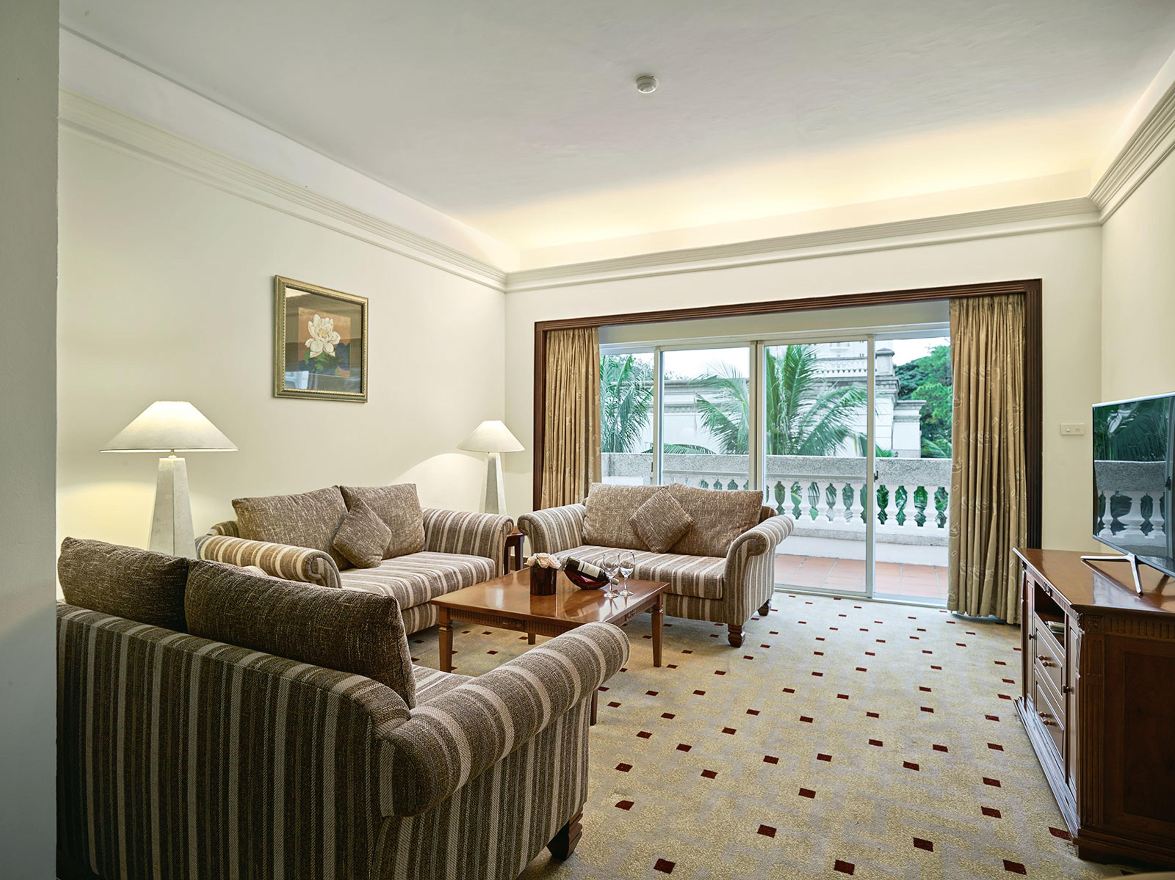 Phòng khách villa với ba ghế sofa vải sọc quanh bàn trà gỗ, hai đèn cây và cửa kính trượt mở ra ban công lan can con tiện