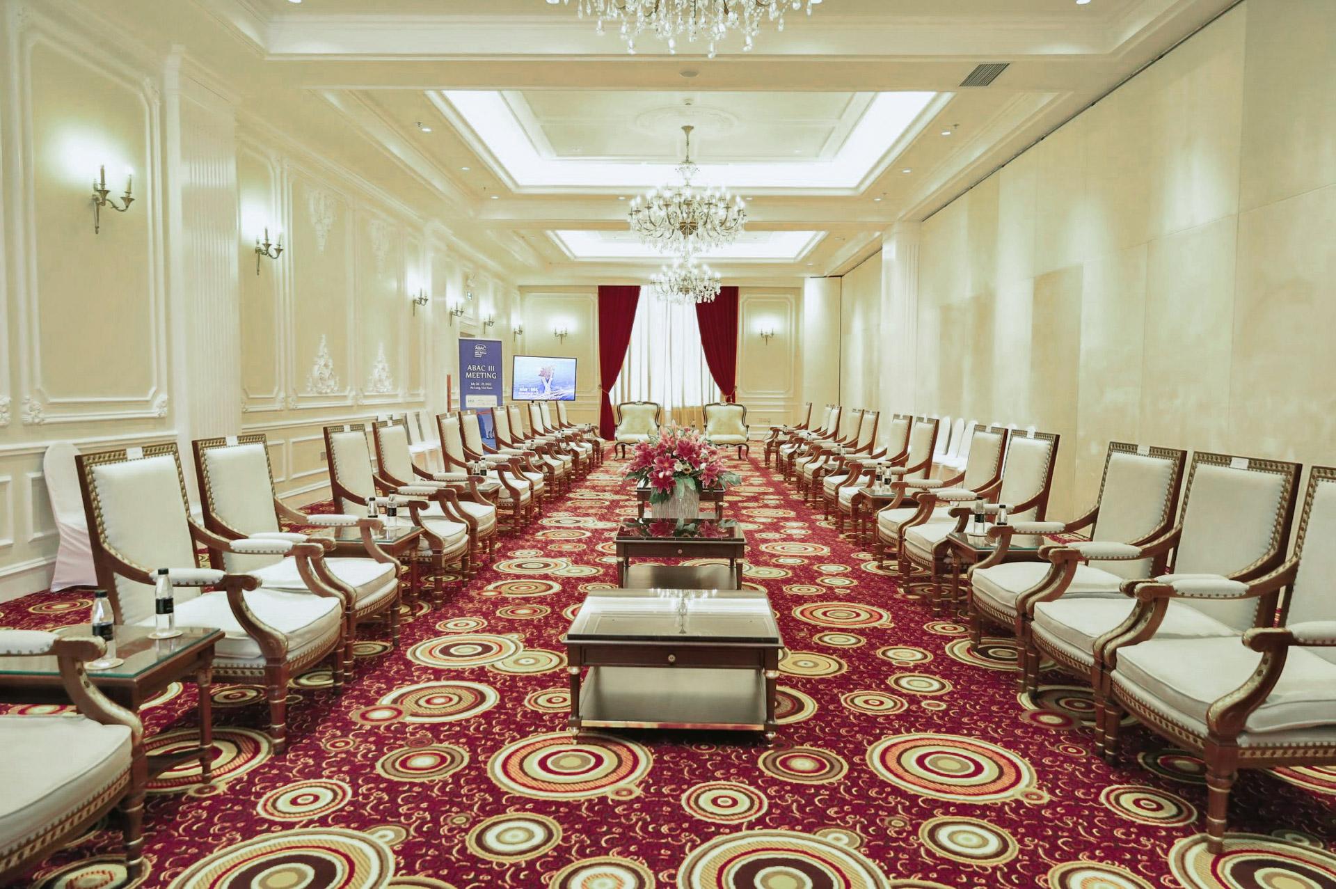 Hoang Gia Ball Room จัดแบบห้องรับรอง เก้าอี้นวมโครงไม้สีขาวสองแถวหันเข้าหากันตามแนวพรมแดงลายดอก โต๊ะกลางเตี้ยอยู่ระหว่างกลาง ท้ายห้องมีจอภาพและผ้าม่านกำมะหยี่สีแดง