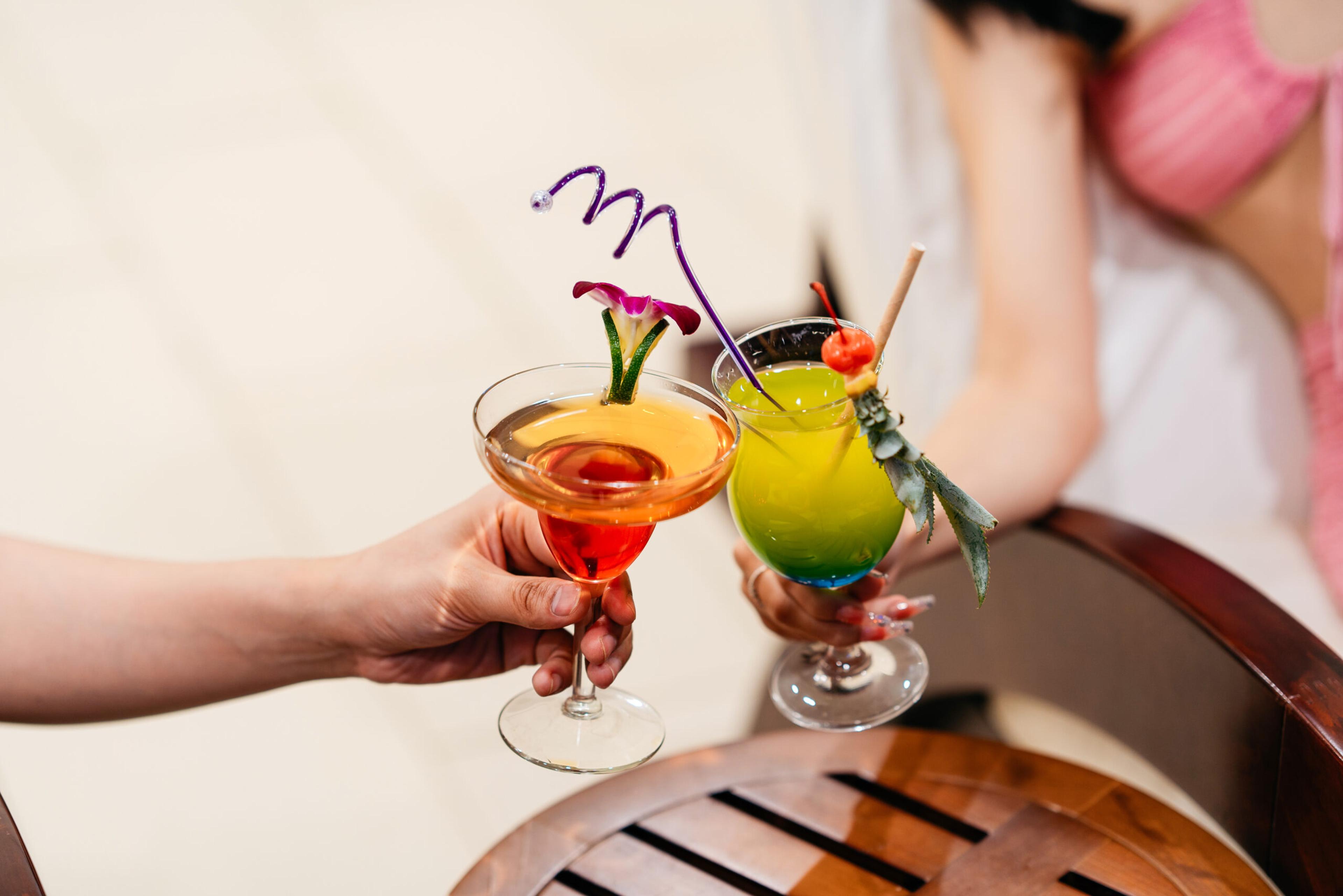 Two hands clinking cocktails — a red martini glass and a tall green one on a wooden tray