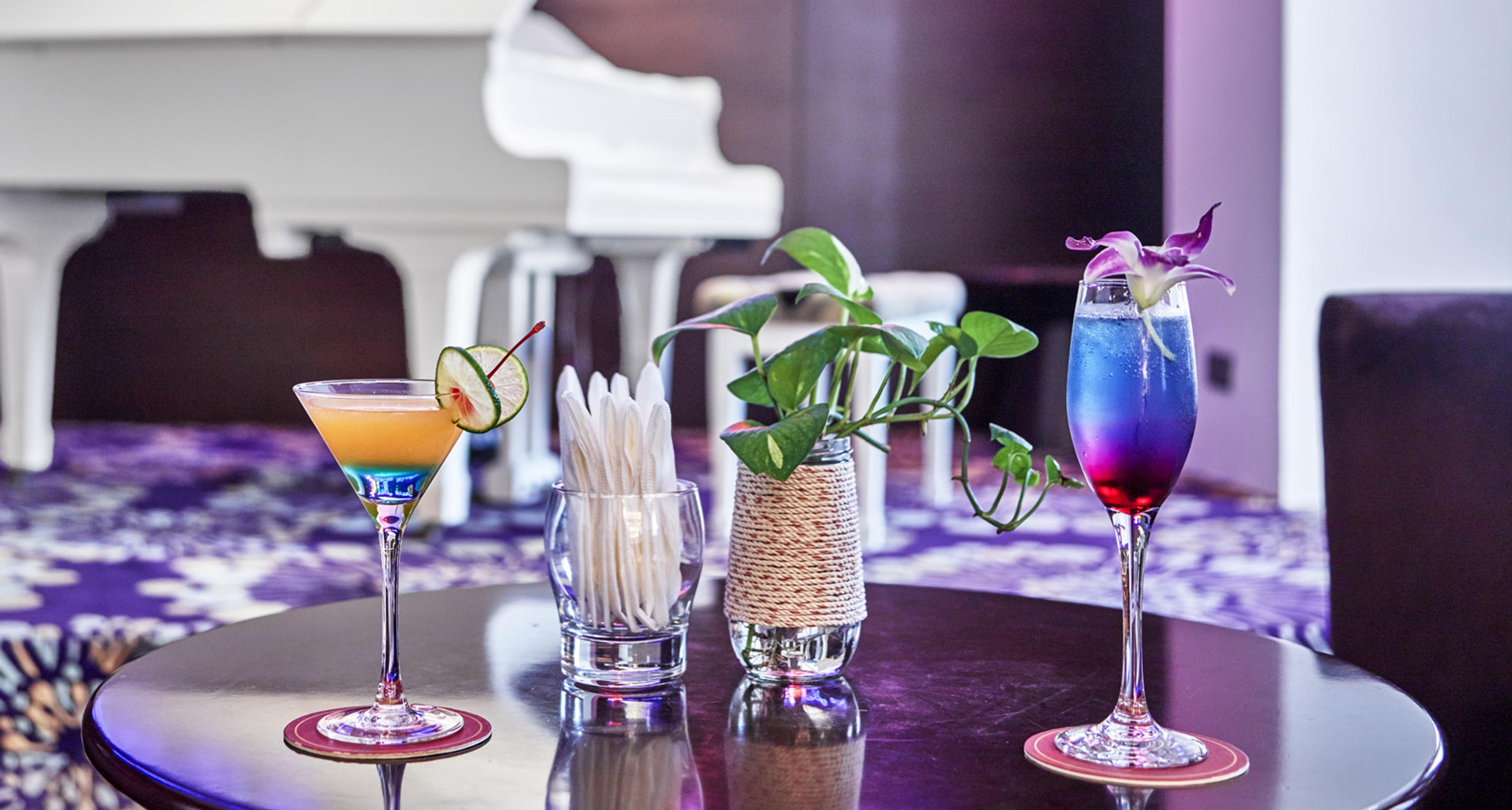 Two colourful cocktails on a round table in front of a white grand piano under purple light