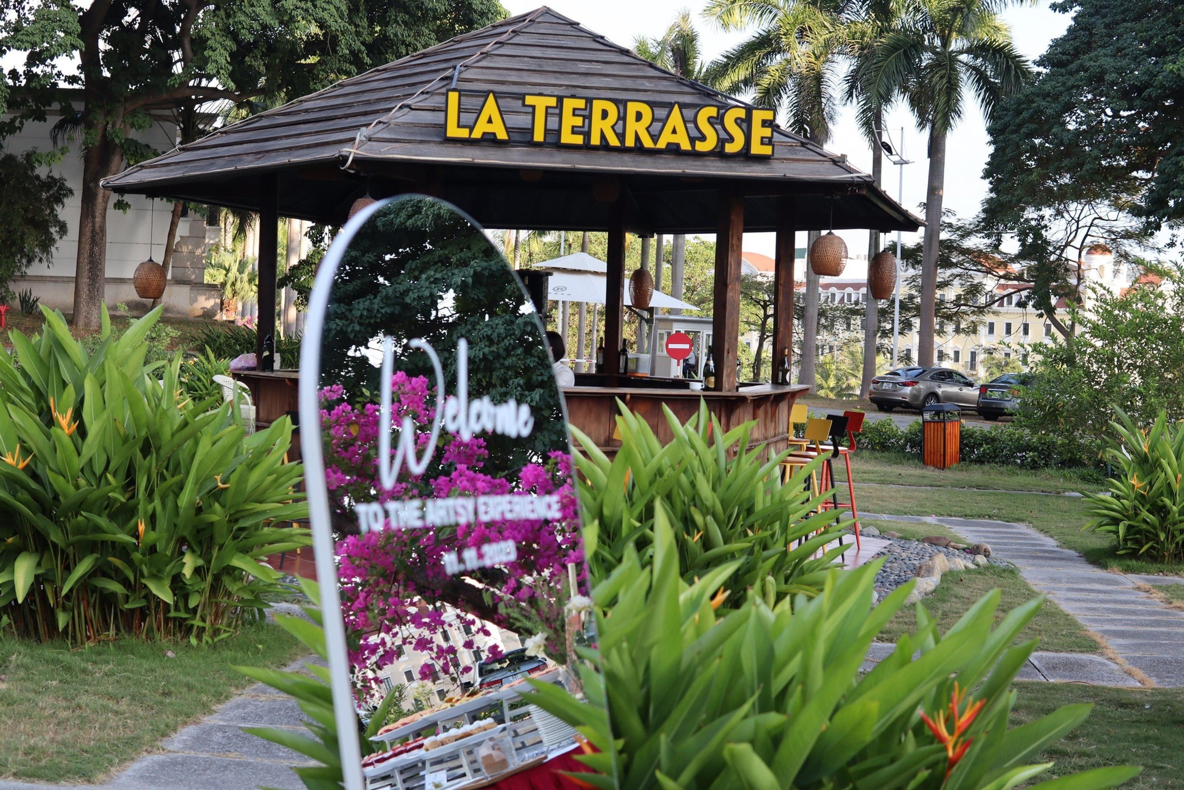 A wooden pavilion carrying the LA TERRASSE sign amid a tropical garden and flower beds, a welcome board beside it