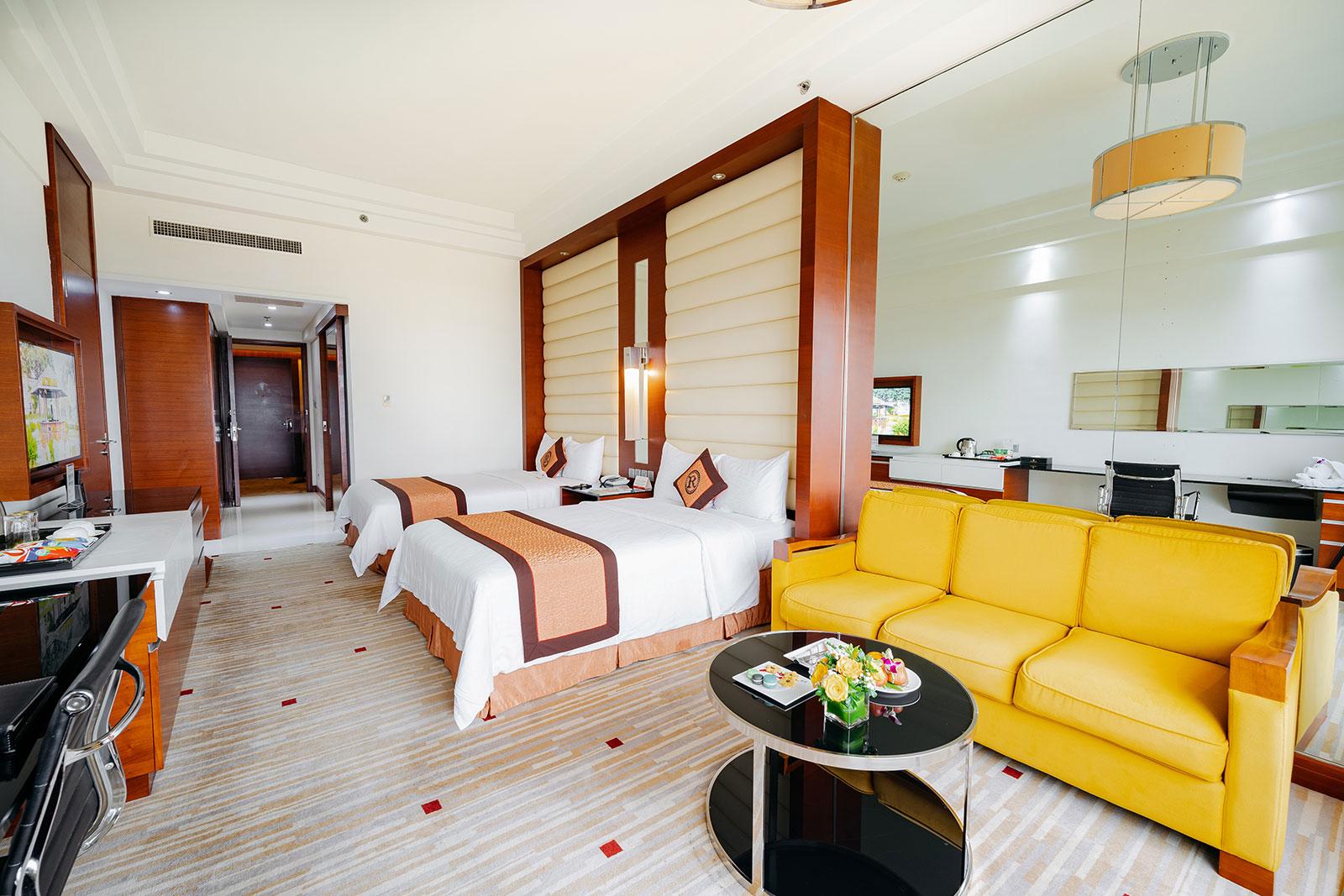 The full Deluxe room: two single beds under orange runners, a yellow sofa with a round glass table, and the timber-panelled entrance on the left