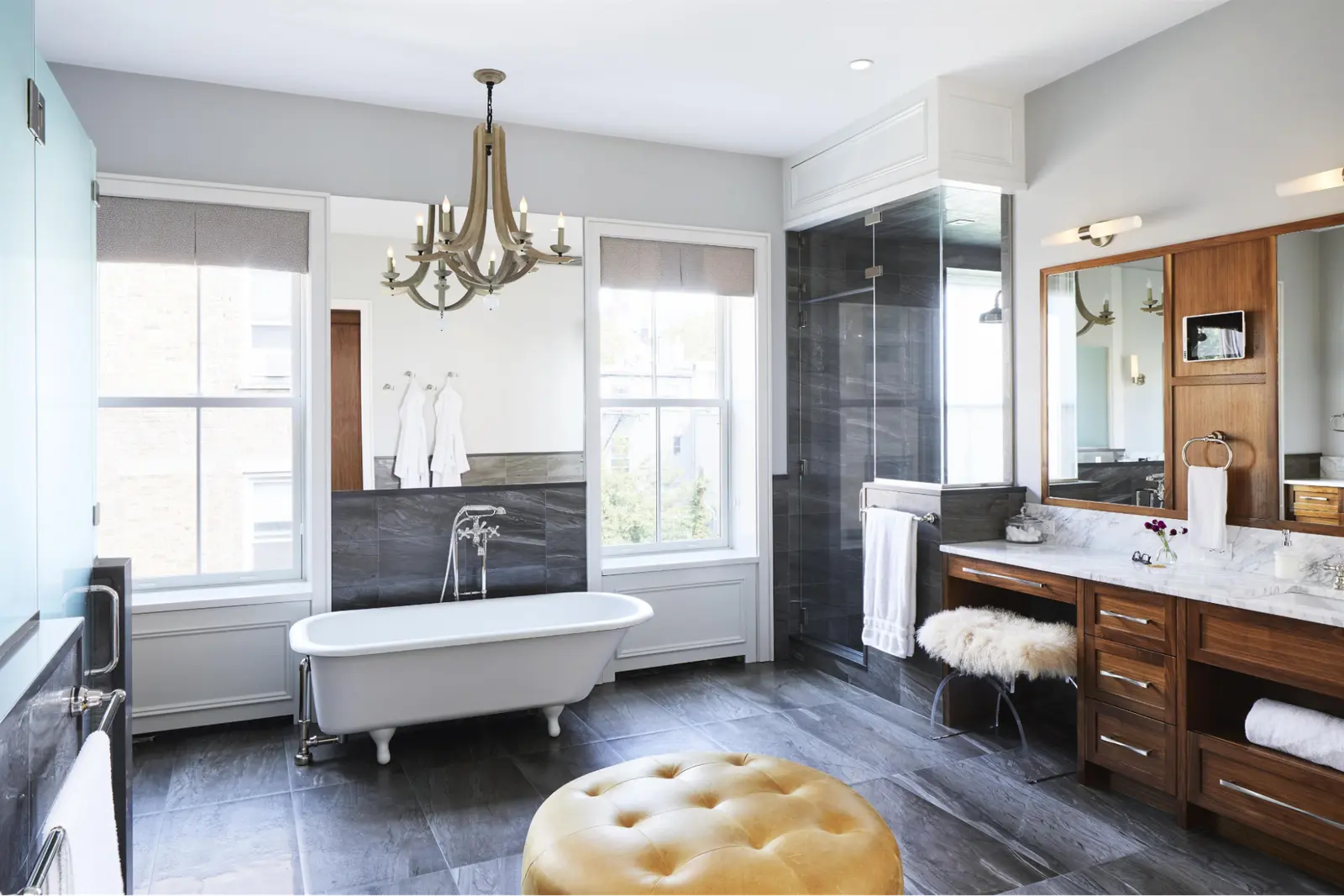 Bright bathroom with a clawfoot tub, chandelier, wooden vanity and a yellow tufted ottoman
