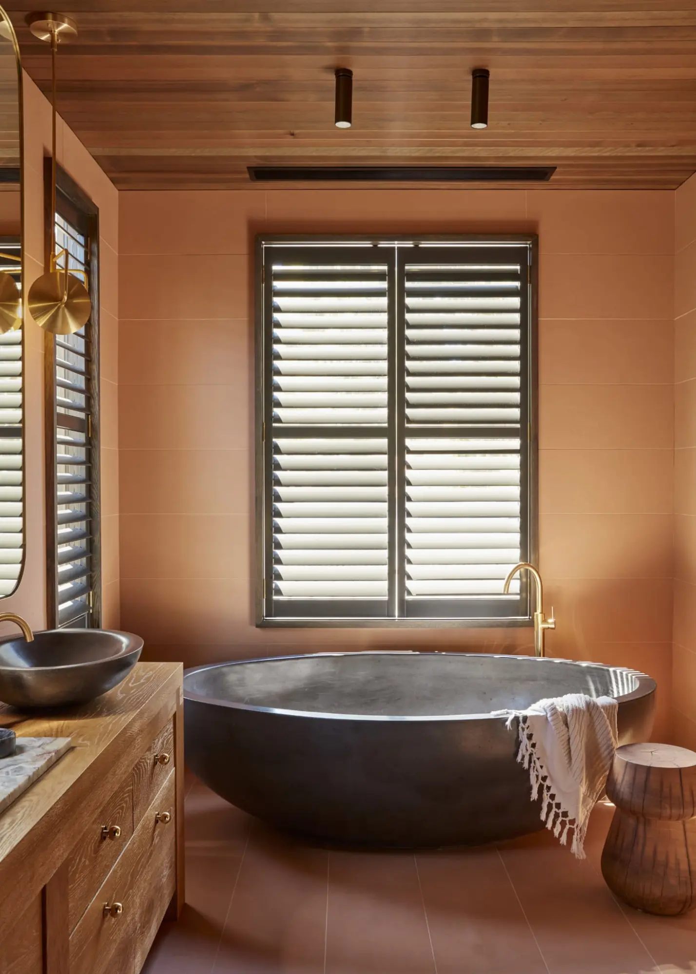 Terracotta bathroom with a dark stone bathtub, wooden vanity and shuttered window