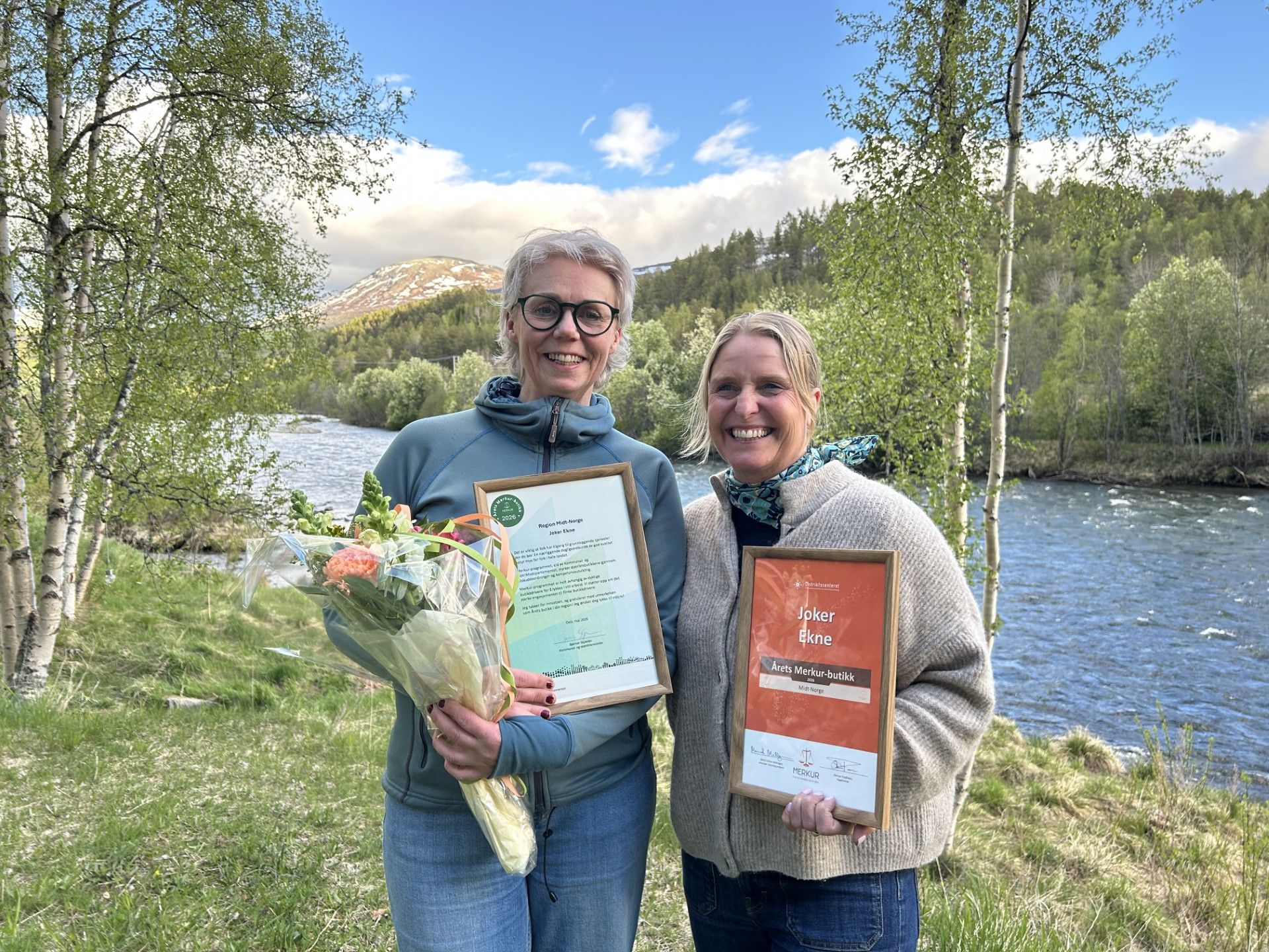 Two smiling women hold framed awards and a bouquet of flowers by a river with a mountainous landscape in the background.