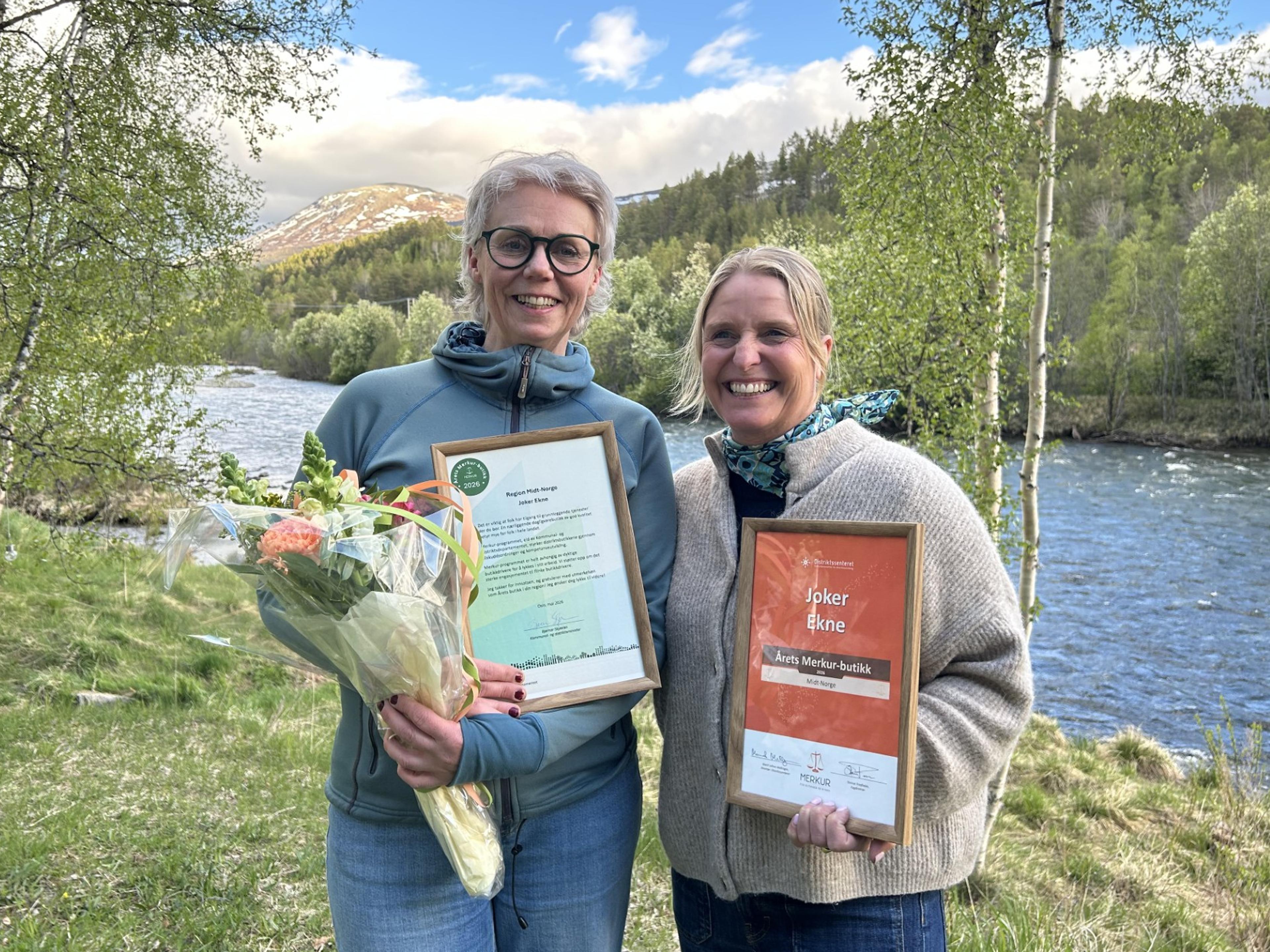 Two smiling women hold framed awards and a bouquet of flowers by a river with a mountainous landscape in the background.