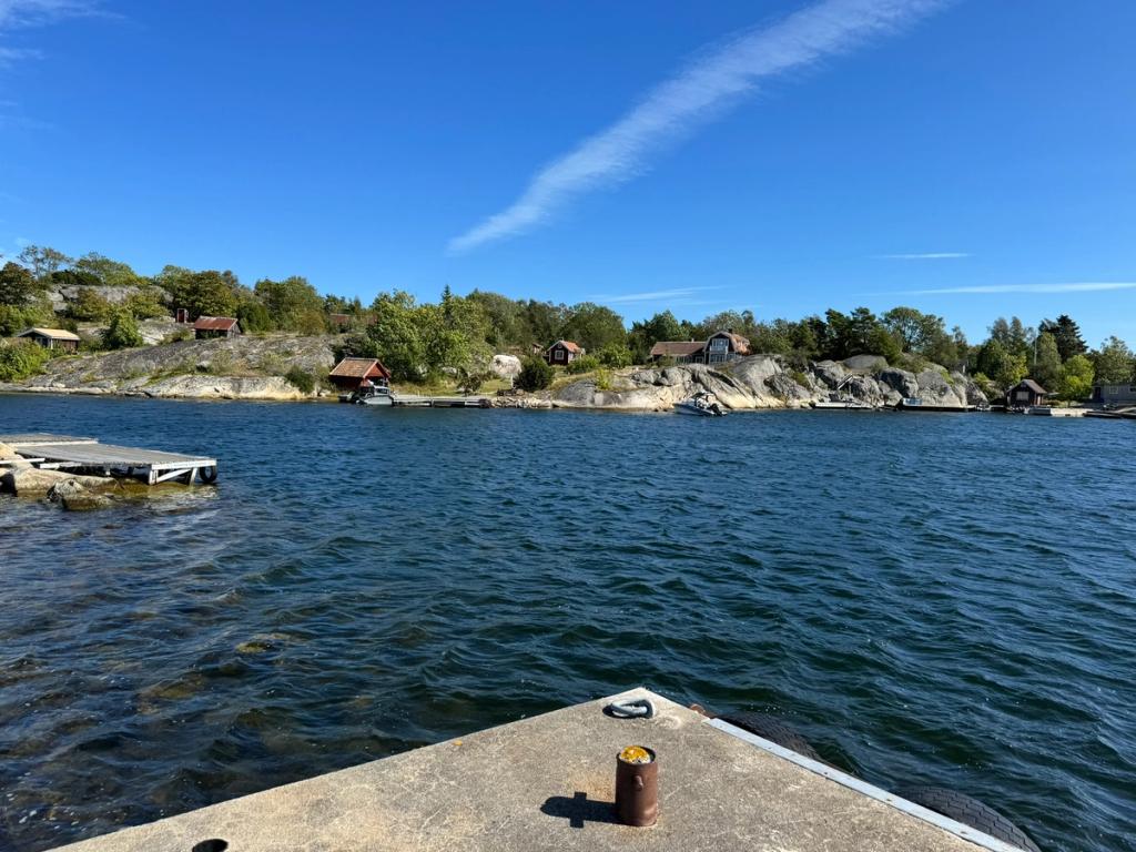 Från en betongbrygga över viken mot strandkanten, där sjöbodar och hus ligger utspridda i skogsbrynet.
