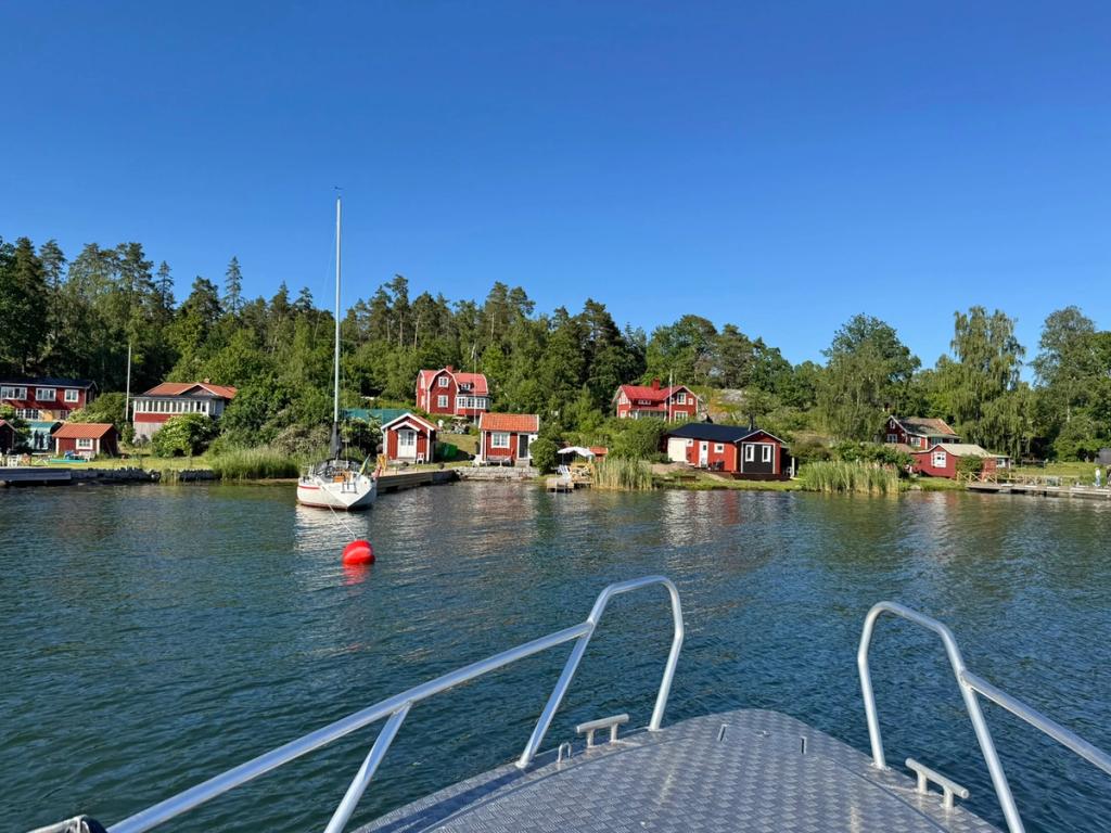 Utsikt från Flanörs för mot en vik med röda stugor, bryggor och en förtöjd segelbåt.