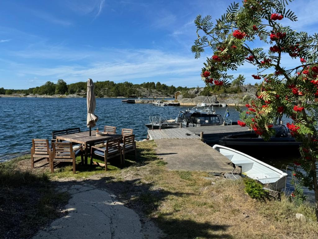 Ett utemöblemang på gräset intill bryggan, med en rönn full av bär och båtar förtöjda bakom.