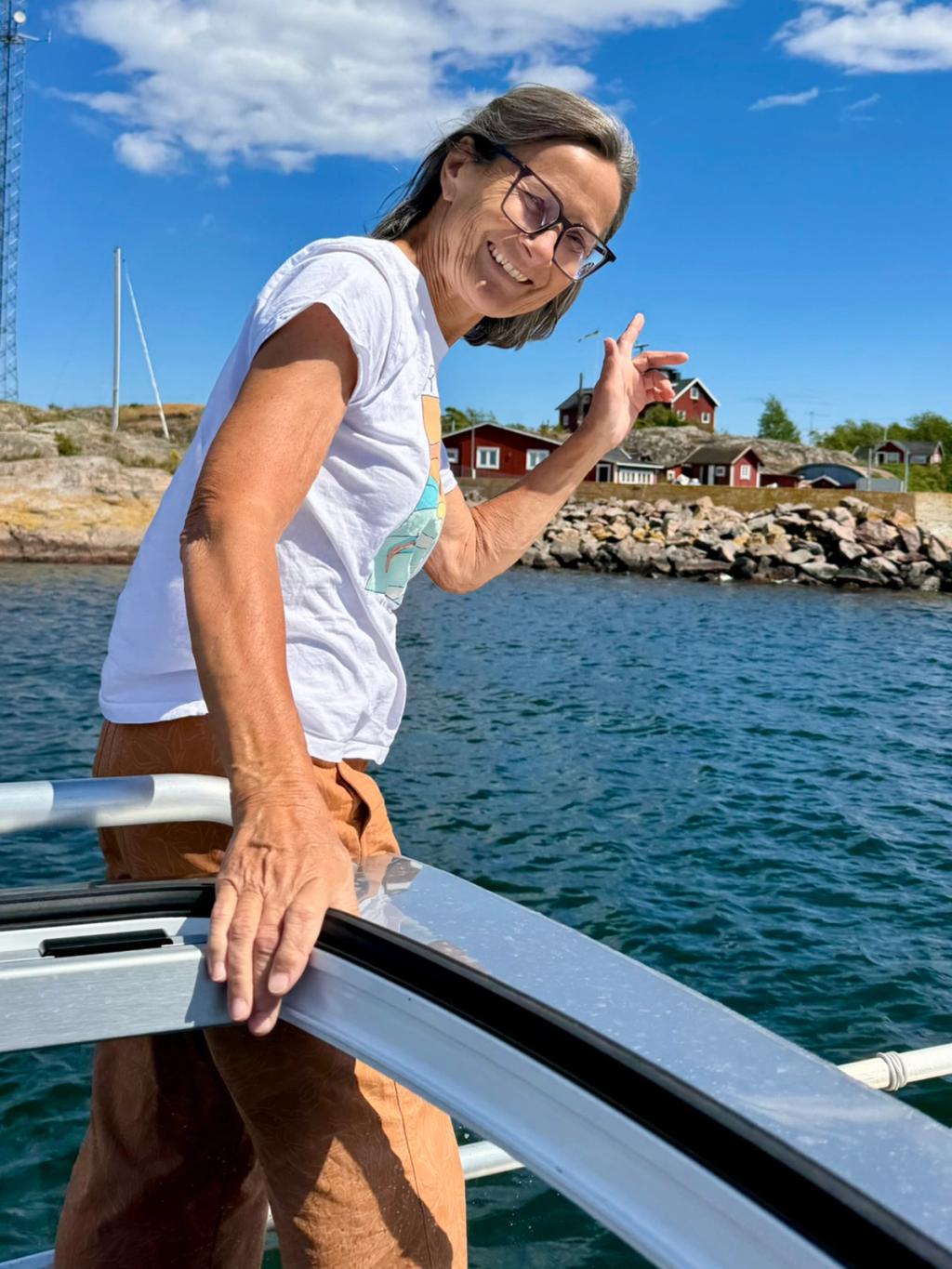 Kari Forsén vinkar från fören, med röda hus och klippor på stranden bakom sig.