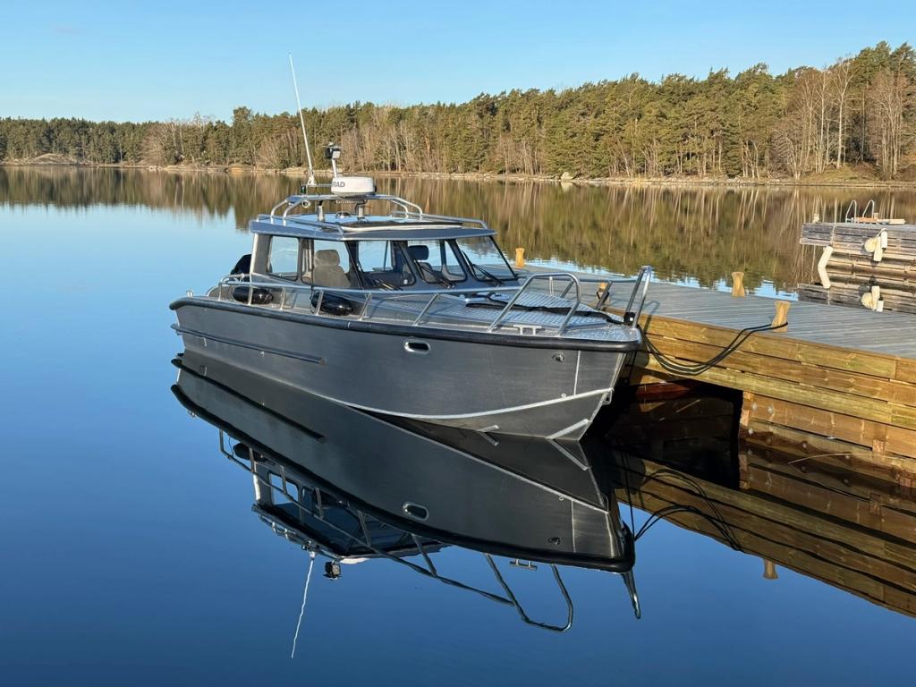 Flanör förtöjd vid en träbrygga på spegelblankt vatten, med skogsklädd strand bakom.