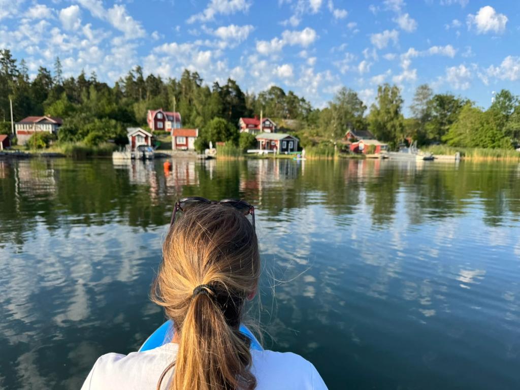En kvinna i fören blickar ut över stilla vatten mot röda hus och bryggor längs stranden.