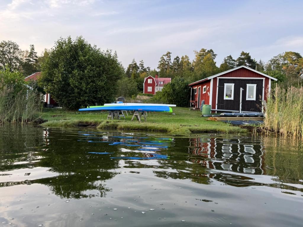 Röd sjöbod och ett rött hus bakom en gräsklädd strand, med kajaker upplagda vid vattenbrynet.