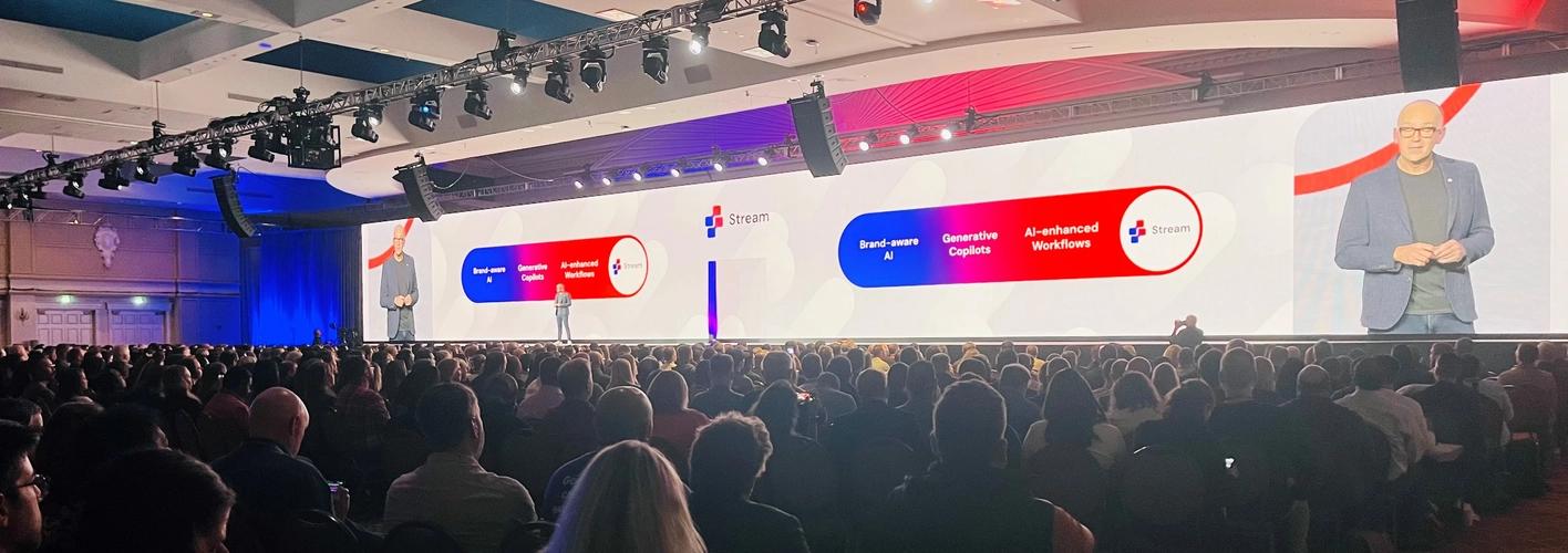 A room full of people watching a presentation about Sitecore Stream with a massive screen behind the speaker amplifying his image. (Web Optimized)