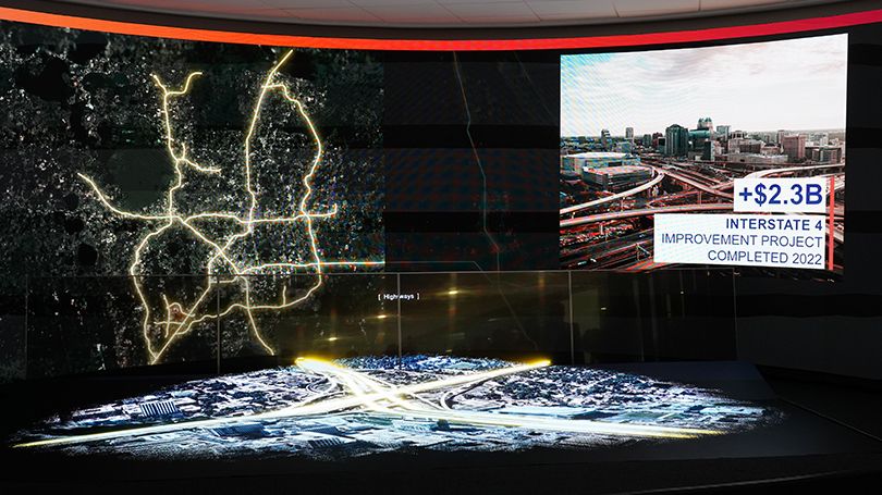The image shows a detailed digital presentation in a dark room, featuring a large curved screen displaying a map with illuminated highway routes outlined in bright lines. Below the map, there is a 3D model of a highway interchange with multiple lanes, ramps, and connecting roads, highlighted with white and yellow lighting effects. In the upper right corner of the screen, there is an inset photo of an actual highway interchange in an urban area, accompanied by text indicating a $2.3 billion improvement project for Interstate 4, completed in 2022.