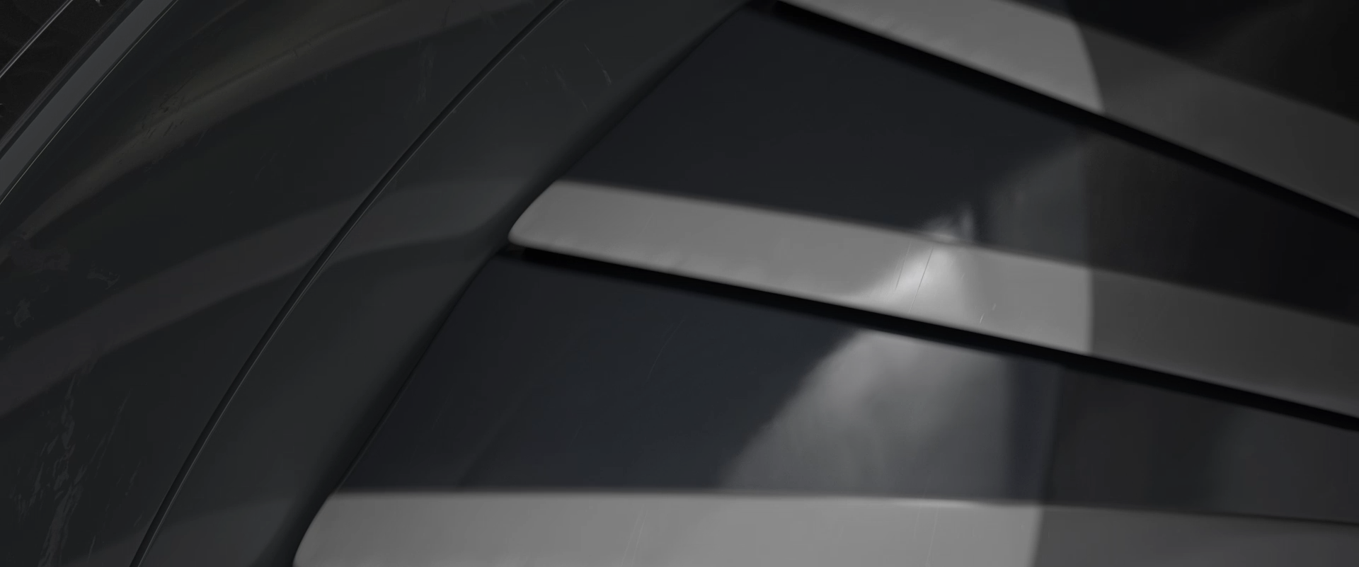 The image shows a close-up view of a dark, glossy surface featuring several parallel, slightly curved metallic slats or fins. The surface reflects light, creating highlights and shadows that emphasize the smooth, polished texture. The overall color palette is monochromatic, with shades of black and gray, suggesting a sleek, modern design likely part of a machine or vehicle component.