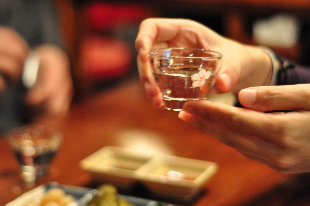 Hai người trao nhau chén soju nhỏ trên bàn nhậu có đồ nhắm